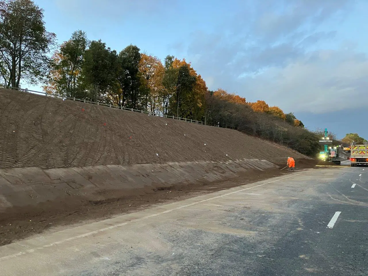 Roadside embankment reinforced with soil stabilization techniques next to a highway. Autumn trees line the top, and construction vehicles and workers in orange safety gear are visible below.