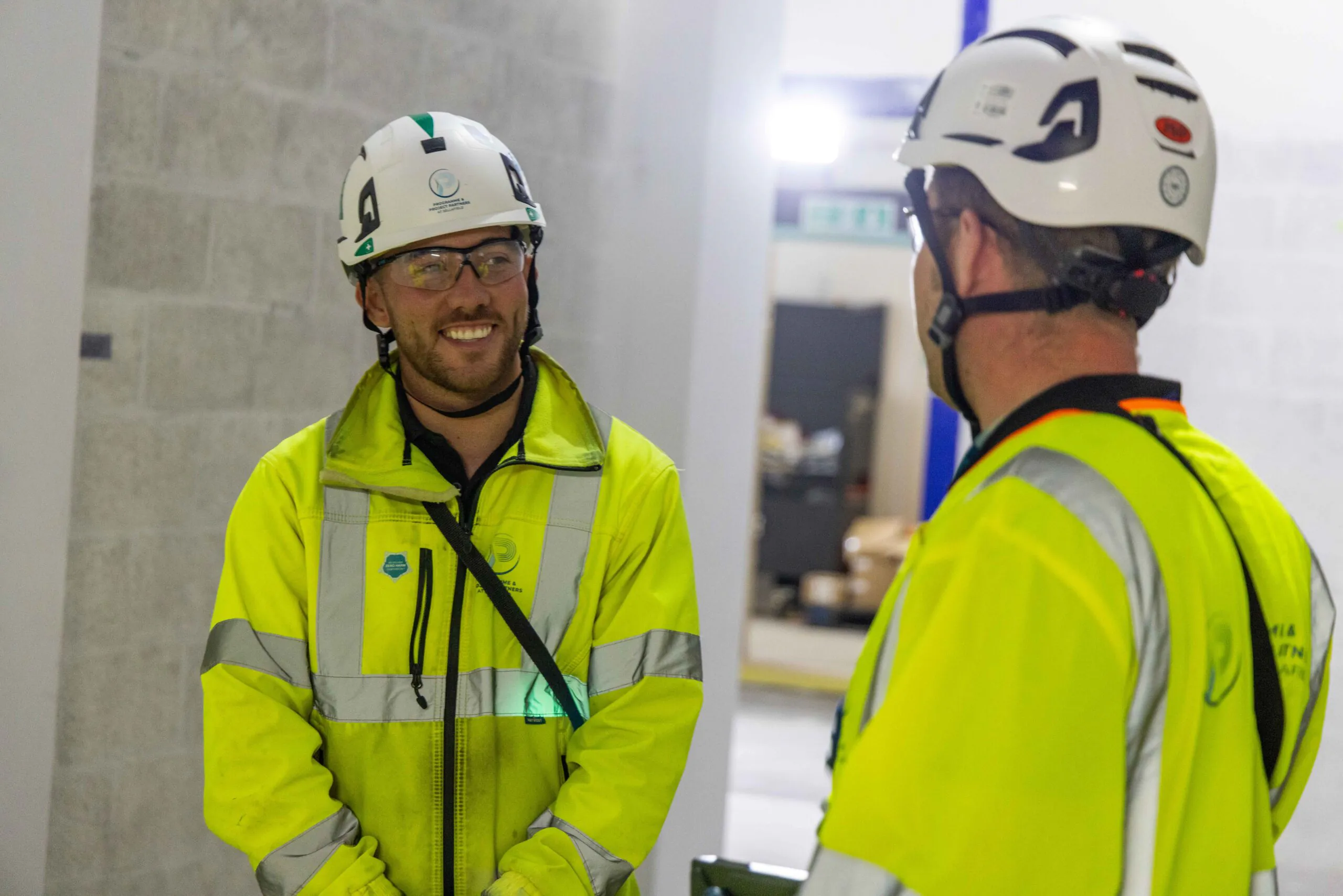 Two construction workers in yellow high-visibility jackets and white helmets converse inside an unfinished building.