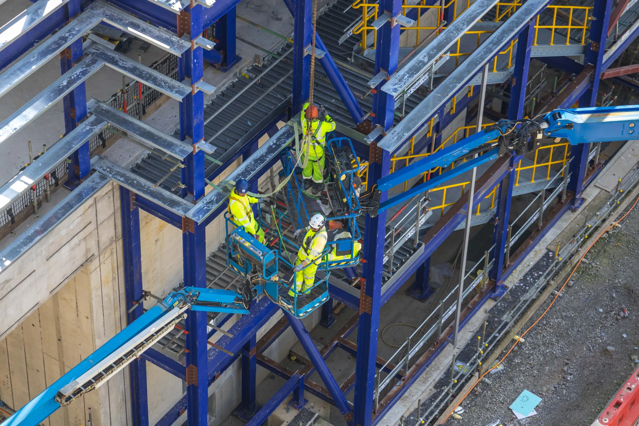 Workers on elevated platforms install or inspect steel framework on an industrial building under construction.