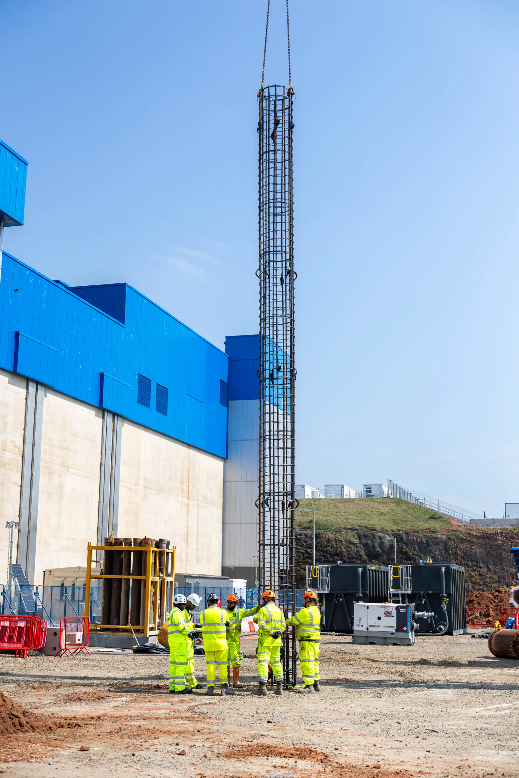 Workers in high-visibility clothing gather around a tall cylindrical structure being lifted by a crane.