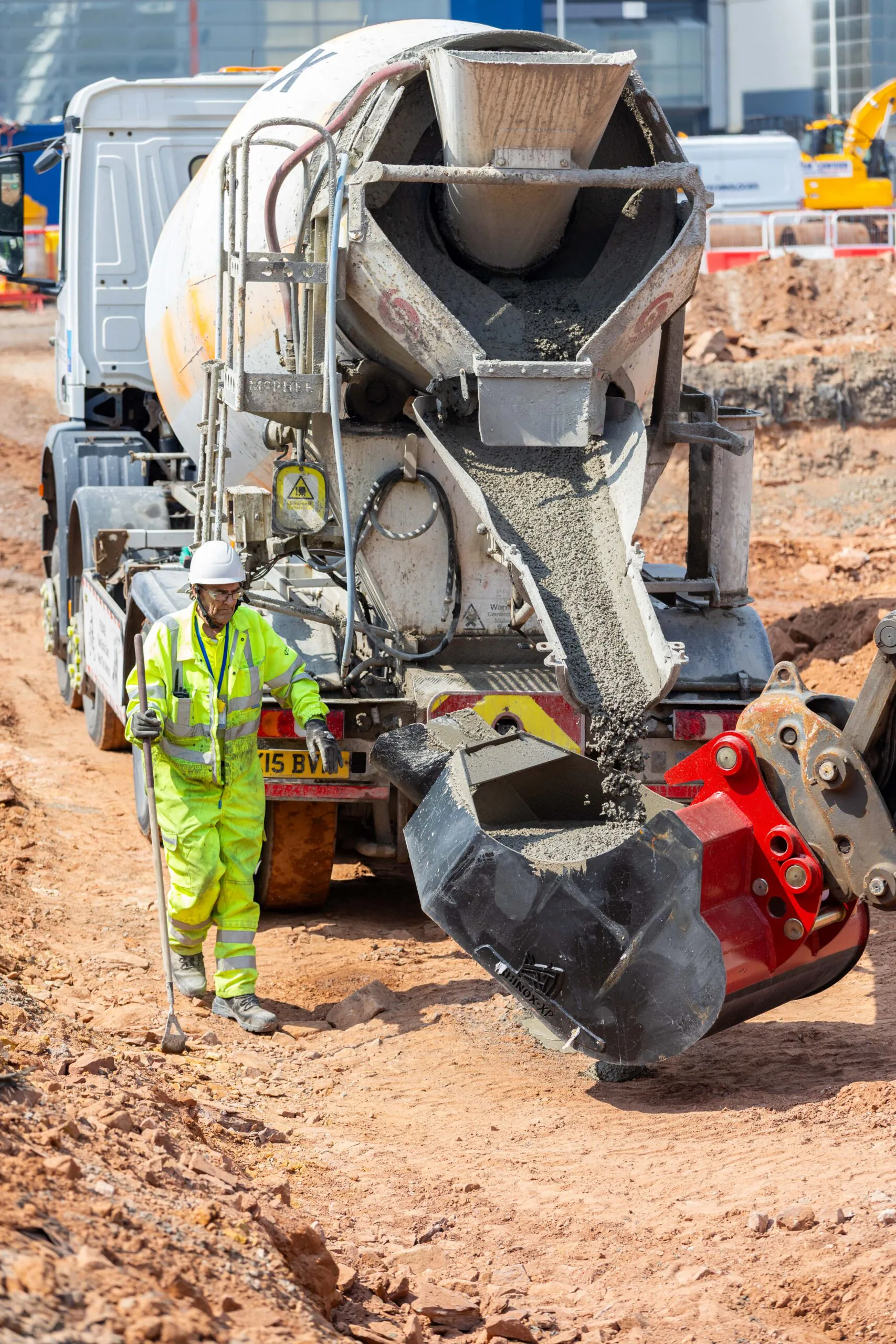 Concrete mixer truck pours concrete into an attachment while a worker in high-visibility clothing stands nearby.