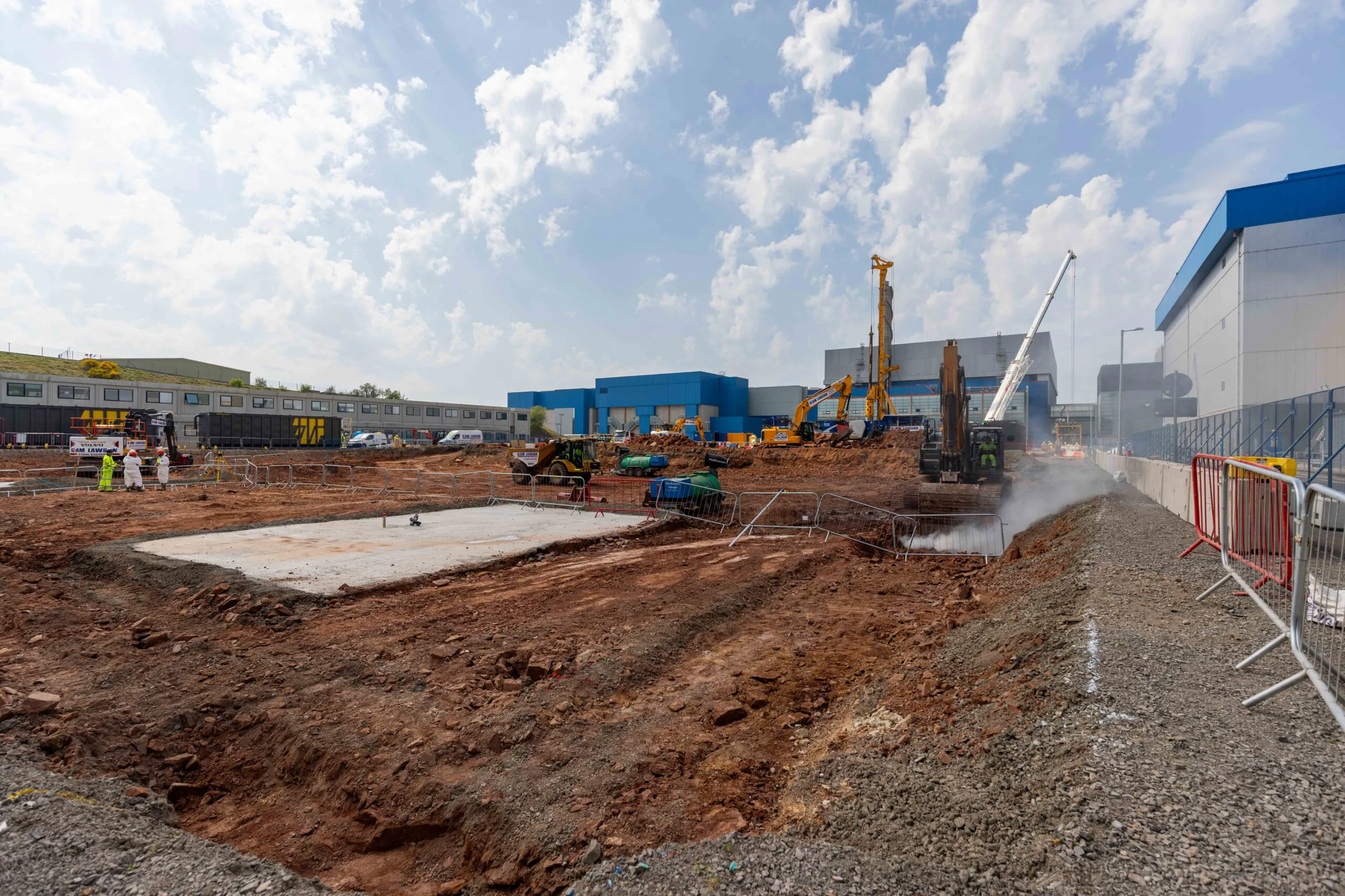 Wide view of an active construction site with cranes, excavators, and surrounding buildings.
