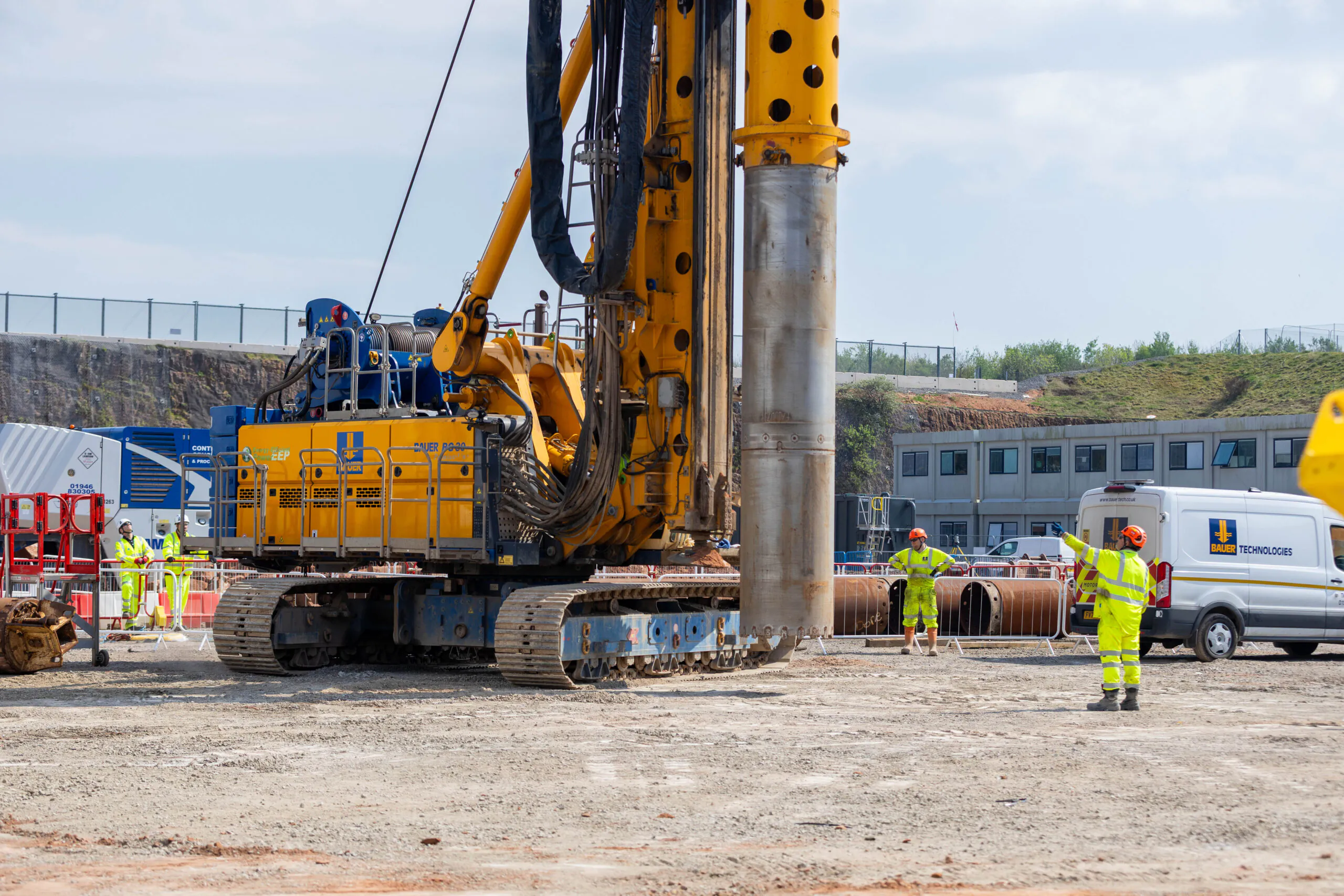 Tall yellow drilling rig operates at a construction site with several workers in high-visibility clothing nearby.