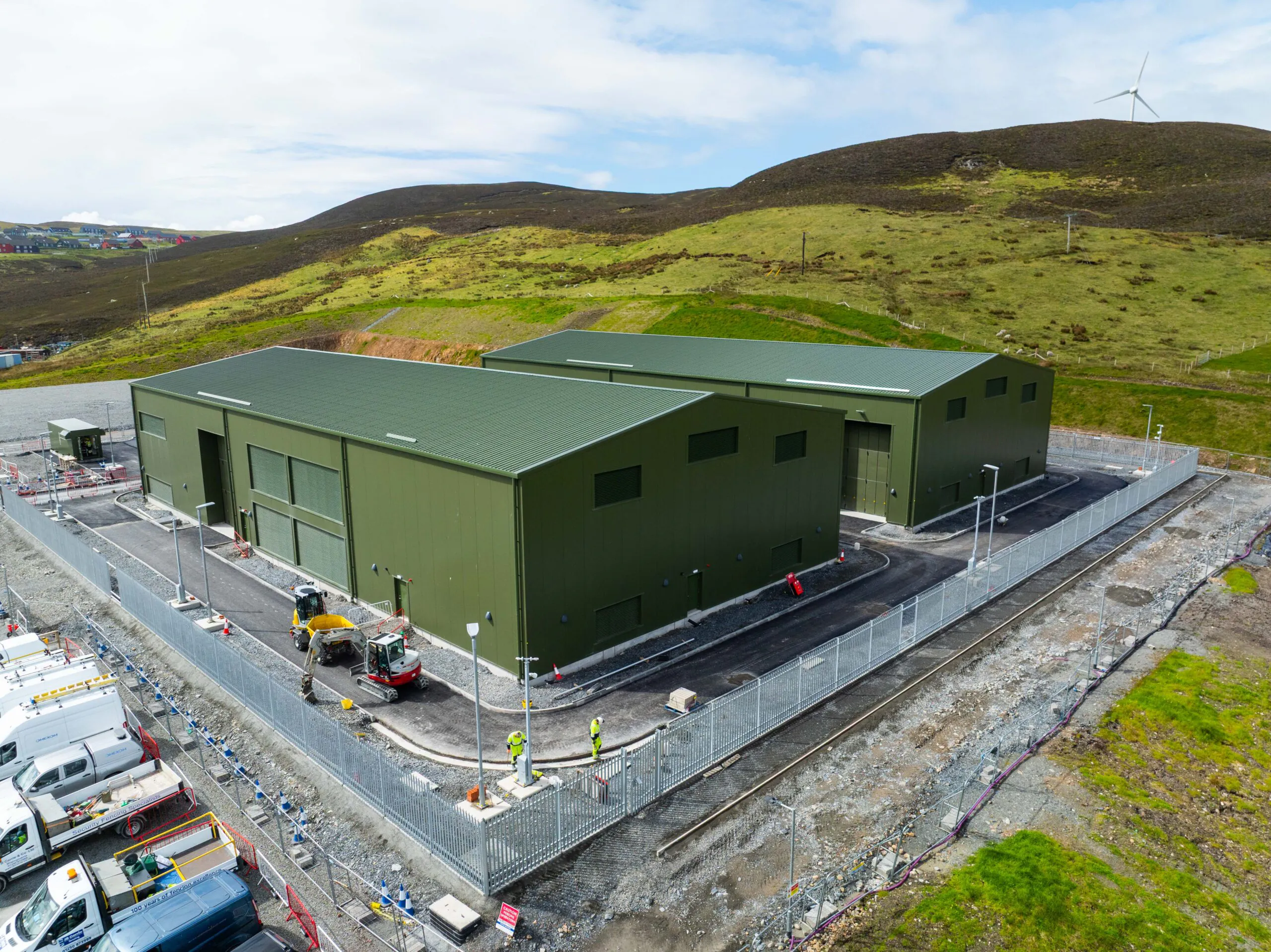 Aerial view of three green industrial buildings surrounded by construction equipment and vehicles within fenced boundaries, set against hilly terrain.
