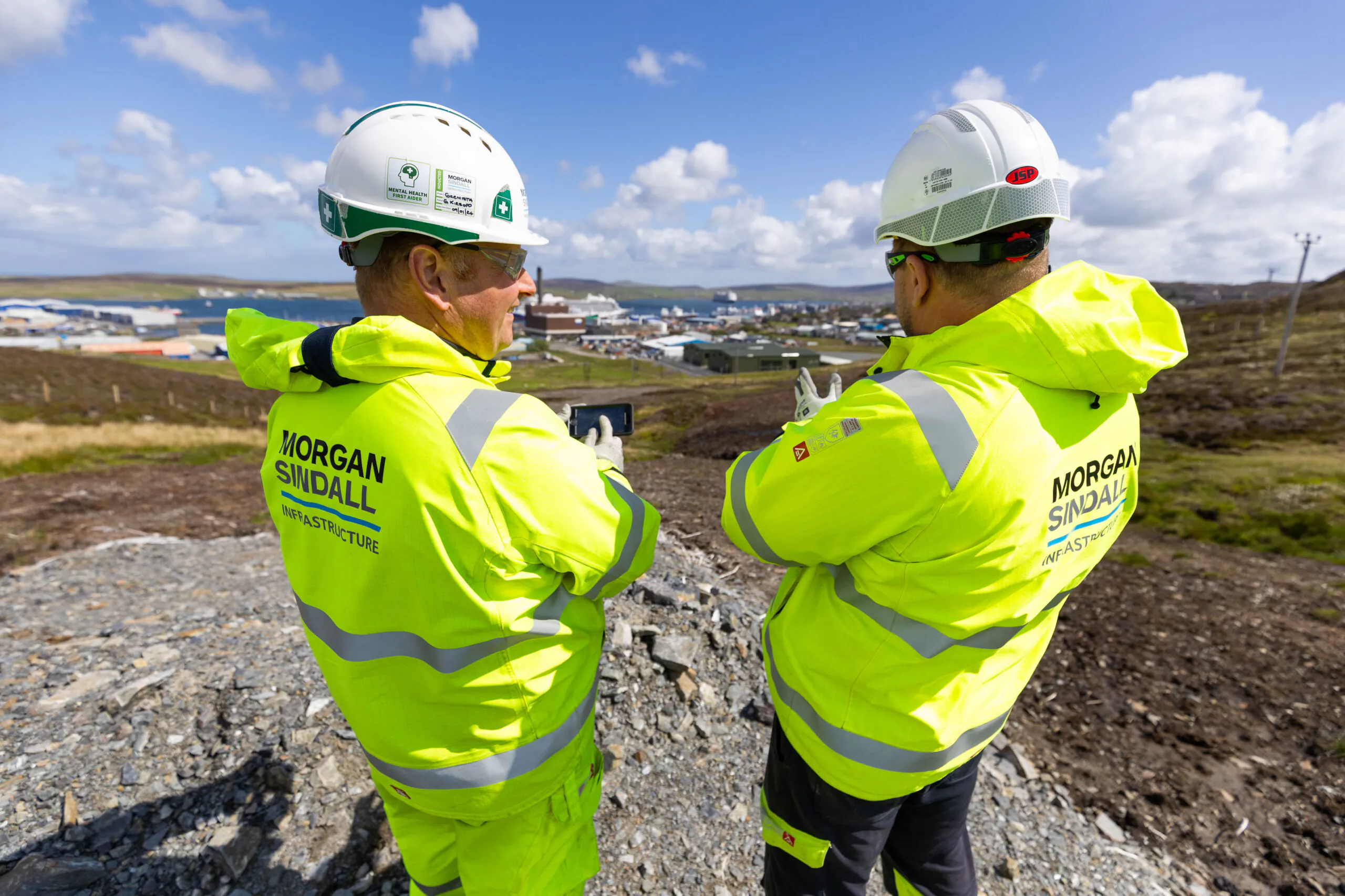 Two individuals in safety gear stand on a rocky hill overlooking a coastal town with docked ships.