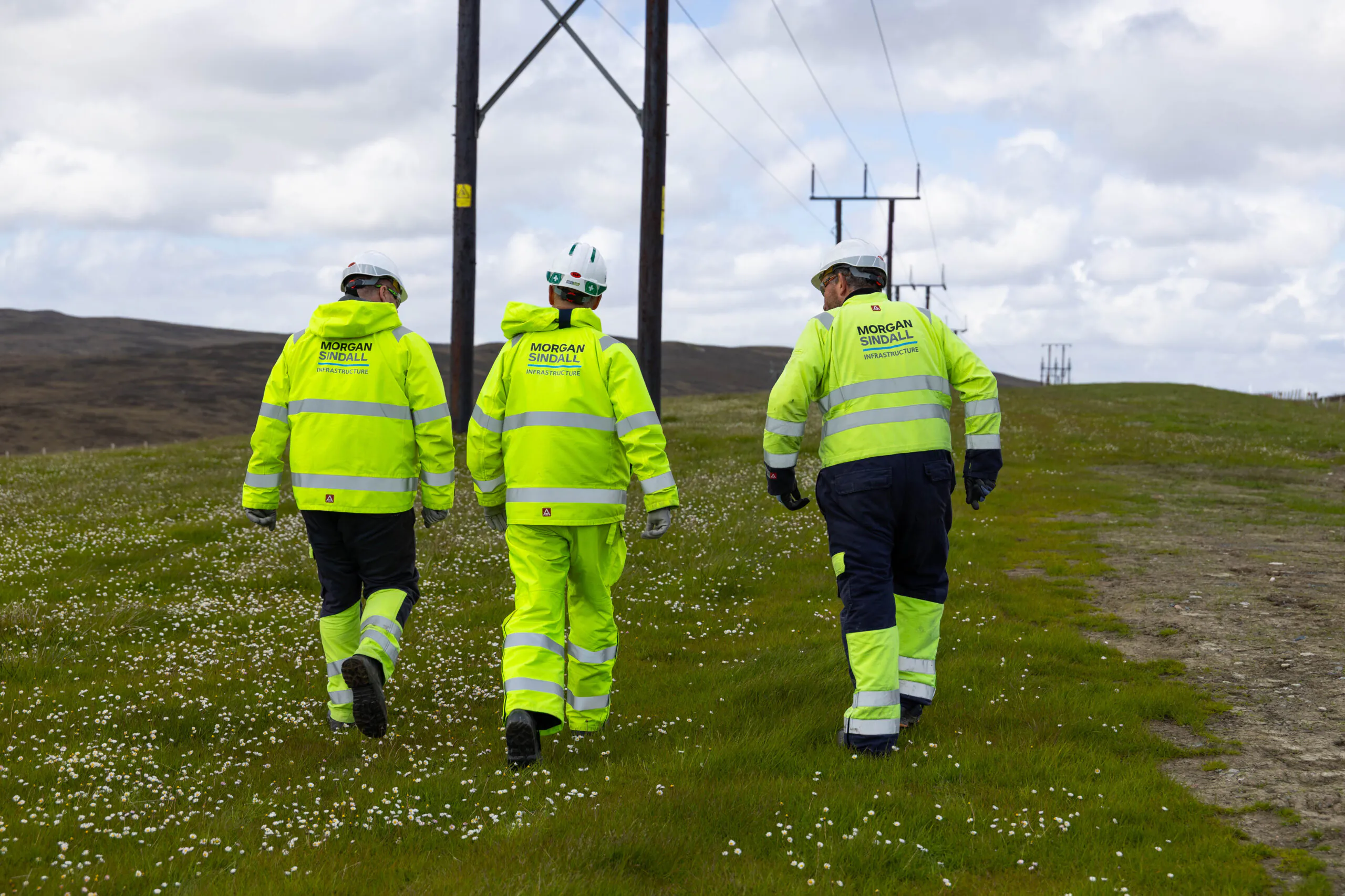 Three individuals in high-visibility jackets labeled "Morgan Sindall" walk along grassy terrain beneath power lines under a partly cloudy sky.