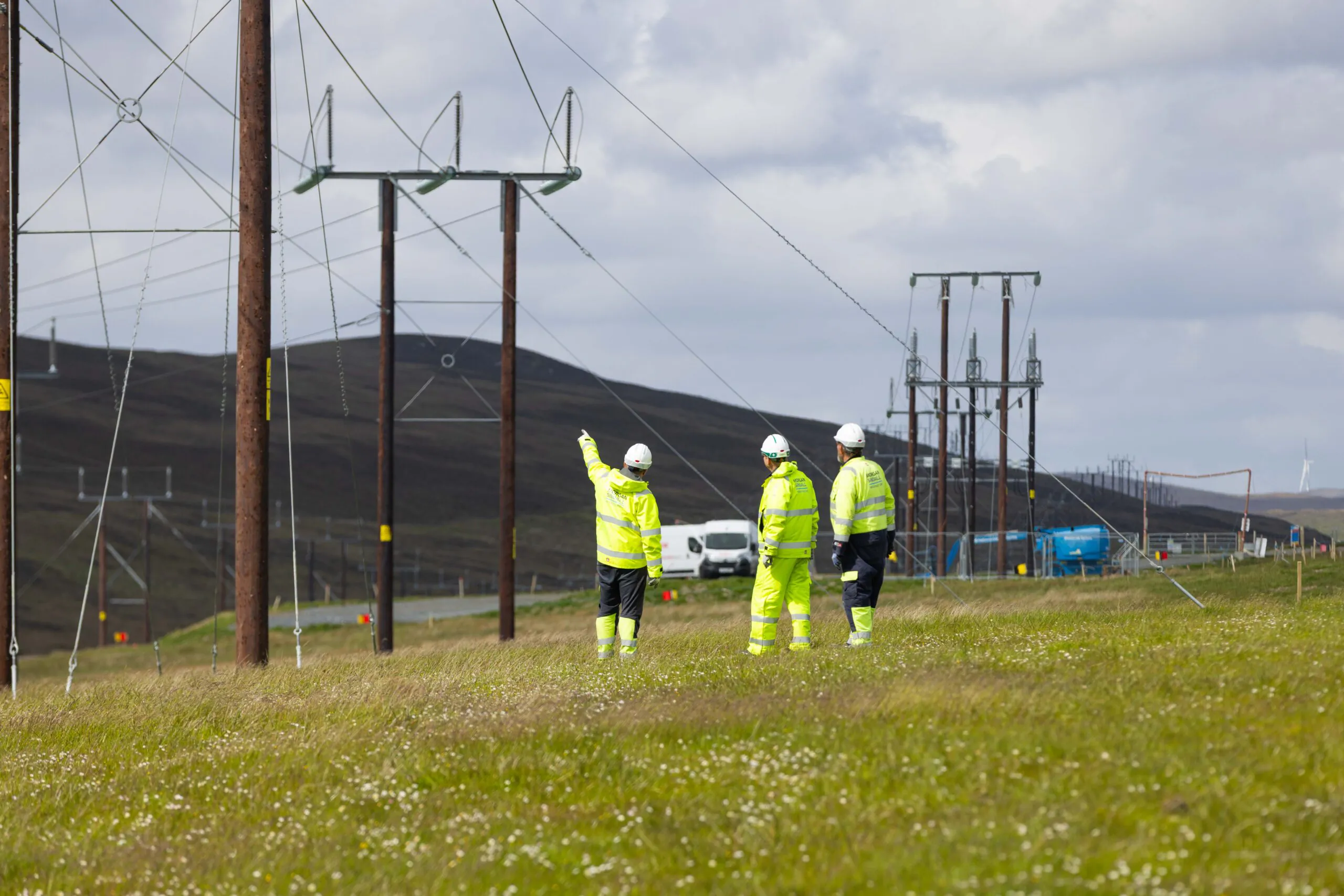 Three individuals in high-visibility jackets labeled "Morgan Sindall" walk along grassy terrain beneath power lines under a partly cloudy sky.