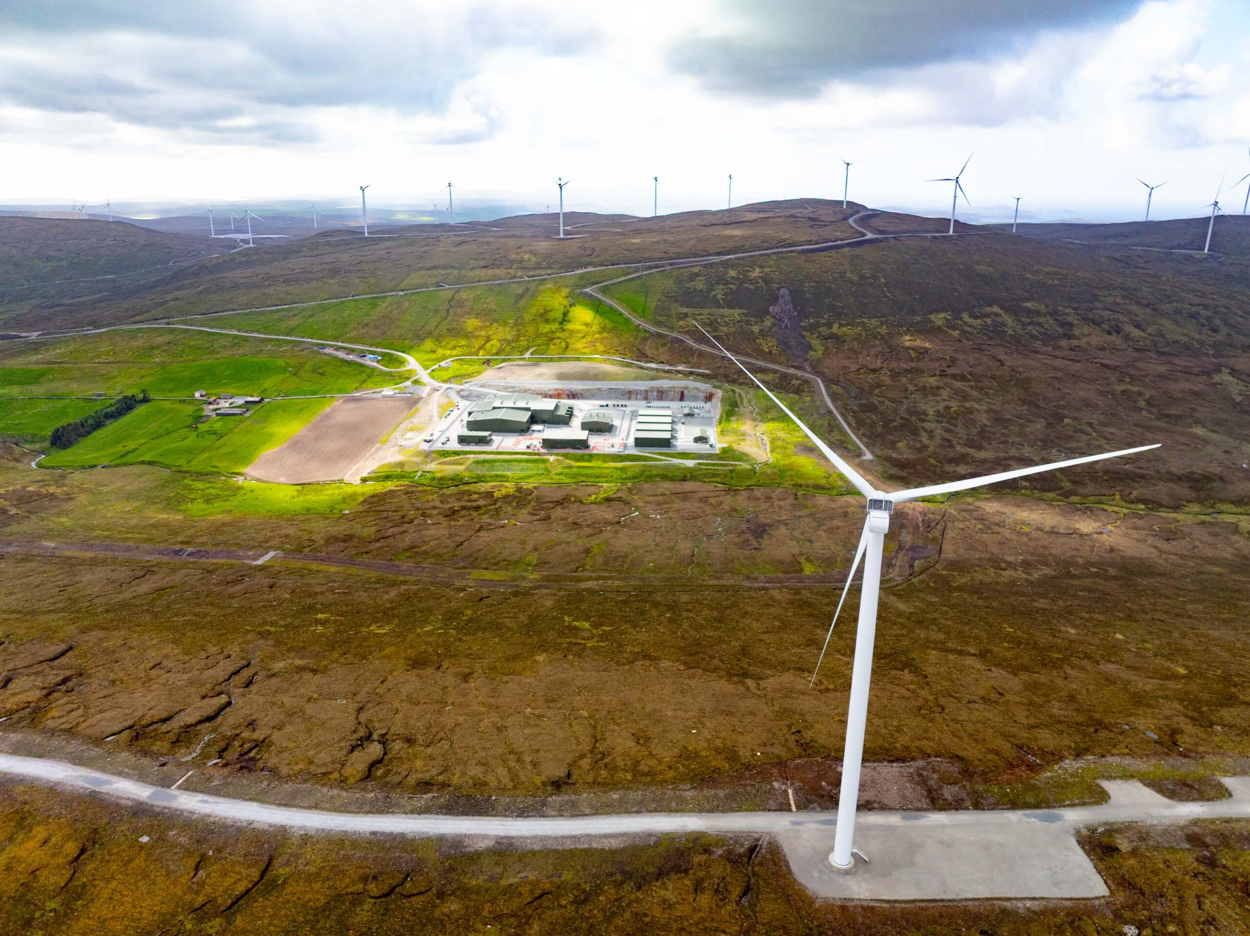 Aerial view of a wind farm with multiple turbines across hilly terrain; a large building complex is visible near the center.