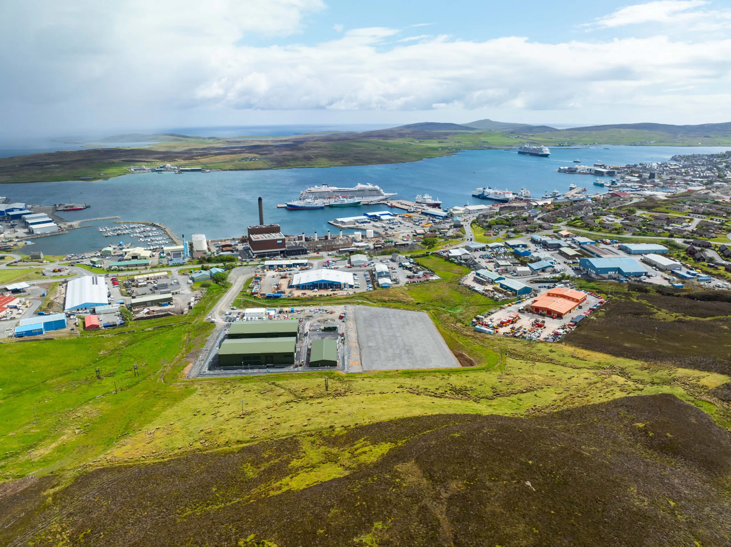 Aerial view of a coastal town with a harbor, showing large ships docked at the port, surrounded by industrial buildings and green hills under a partly cloudy sky.