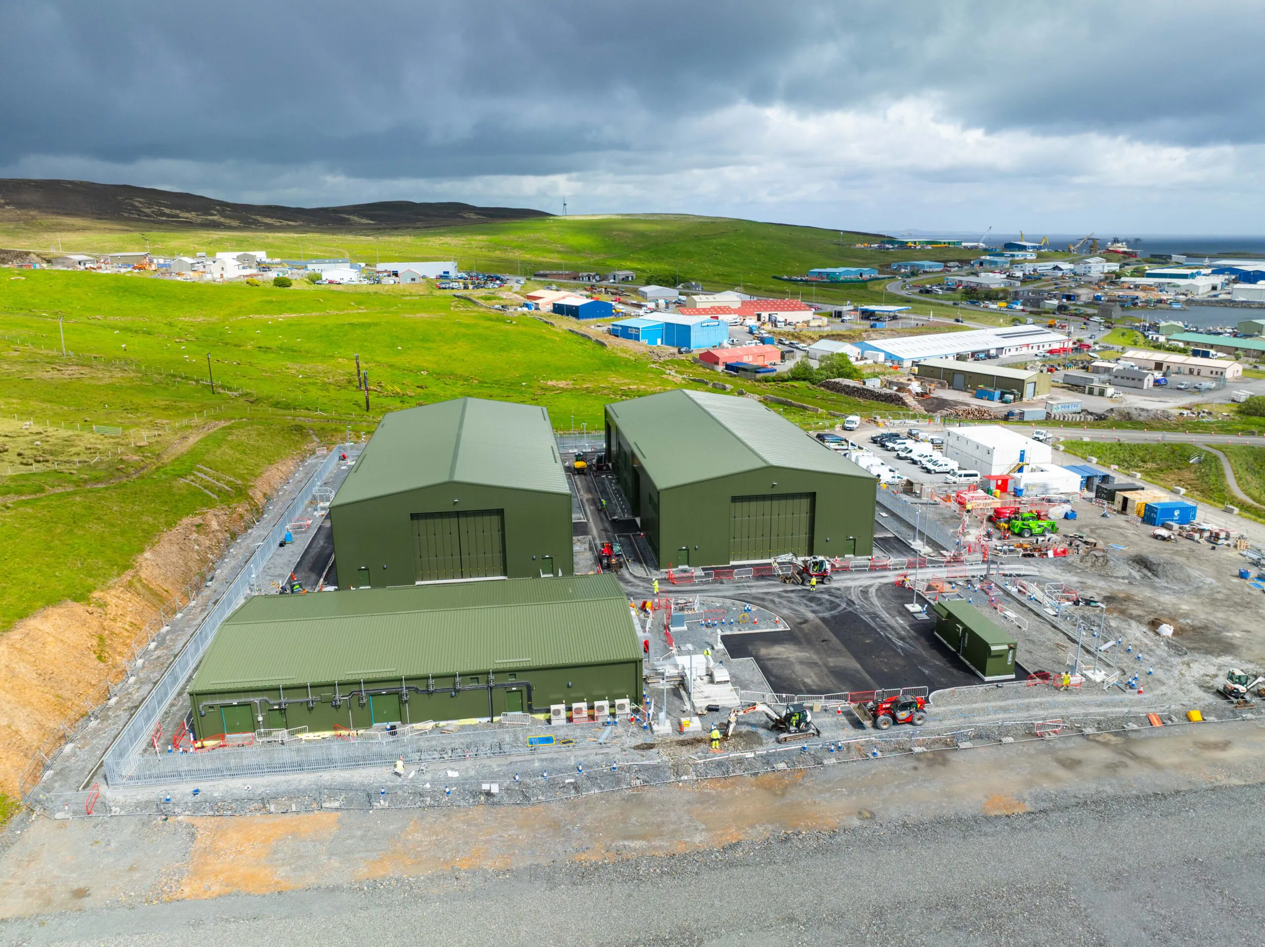 Aerial view of three green industrial buildings surrounded by construction equipment and vehicles within fenced boundaries, set against hilly terrain.