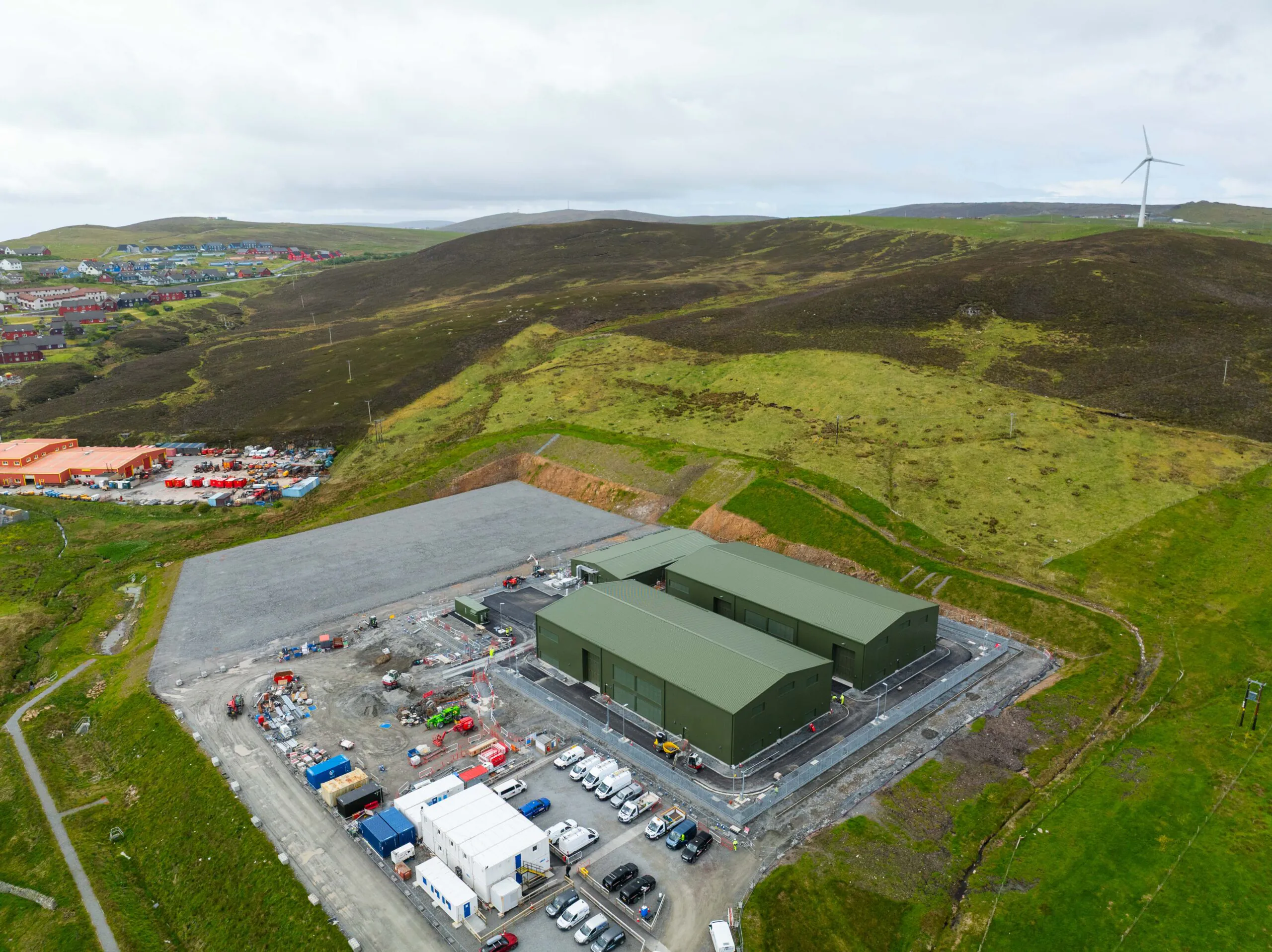 Aerial view of two large green warehouse-like structures surrounded by vehicles and temporary buildings, with wind turbines in the distance.