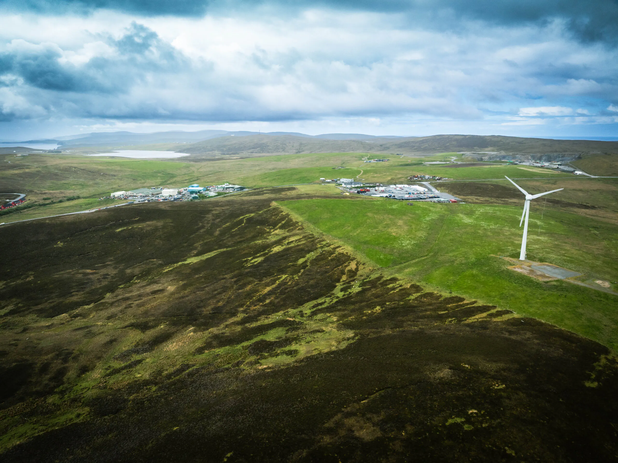 Aerial view of a wind turbine on green hills with an industrial area in the distance under cloudy skies.