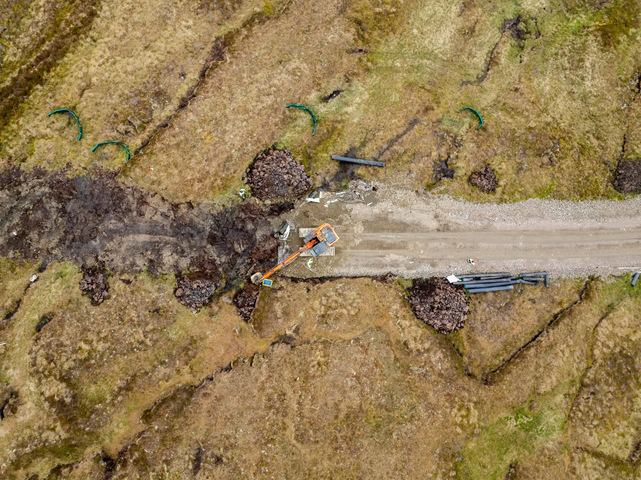 Aerial view of a rural construction site with an excavator working on a dirt road, surrounded by grassy patches, soil, and scattered construction materials.