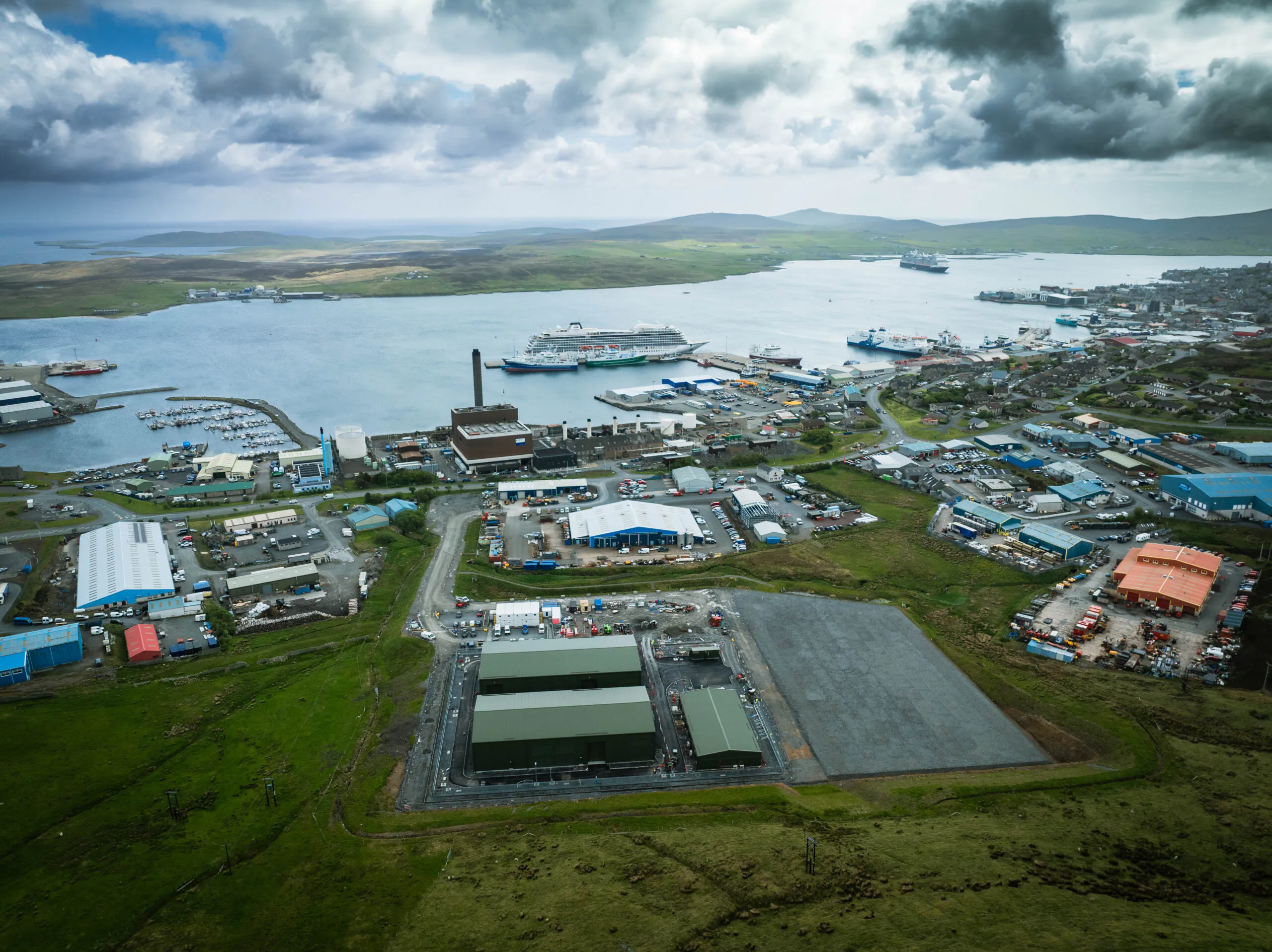Aerial view of an industrial port with buildings, ships docked at piers, and surrounding water, fields, and hills.