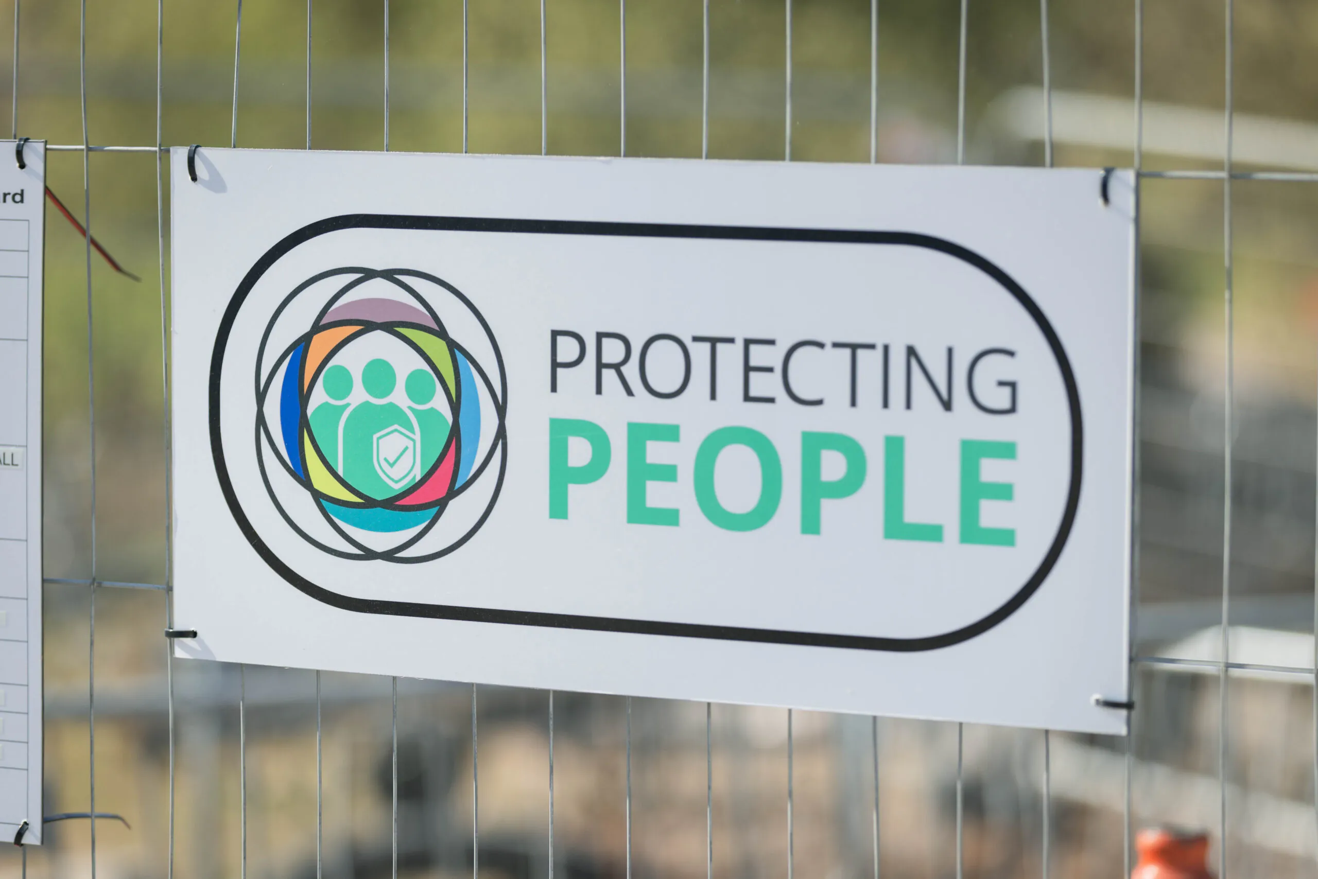 A sign on fencing reads “Protecting People” with colorful icons representing individuals.