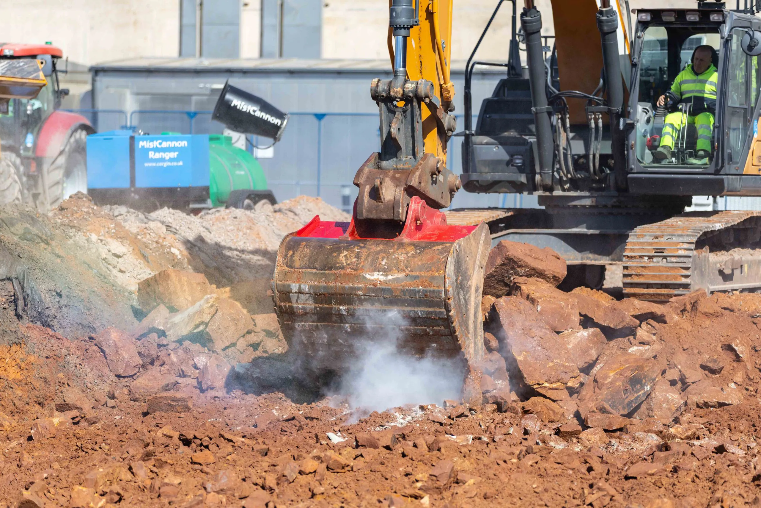 Excavator loads soil into another machine’s bucket at a construction site near industrial buildings.