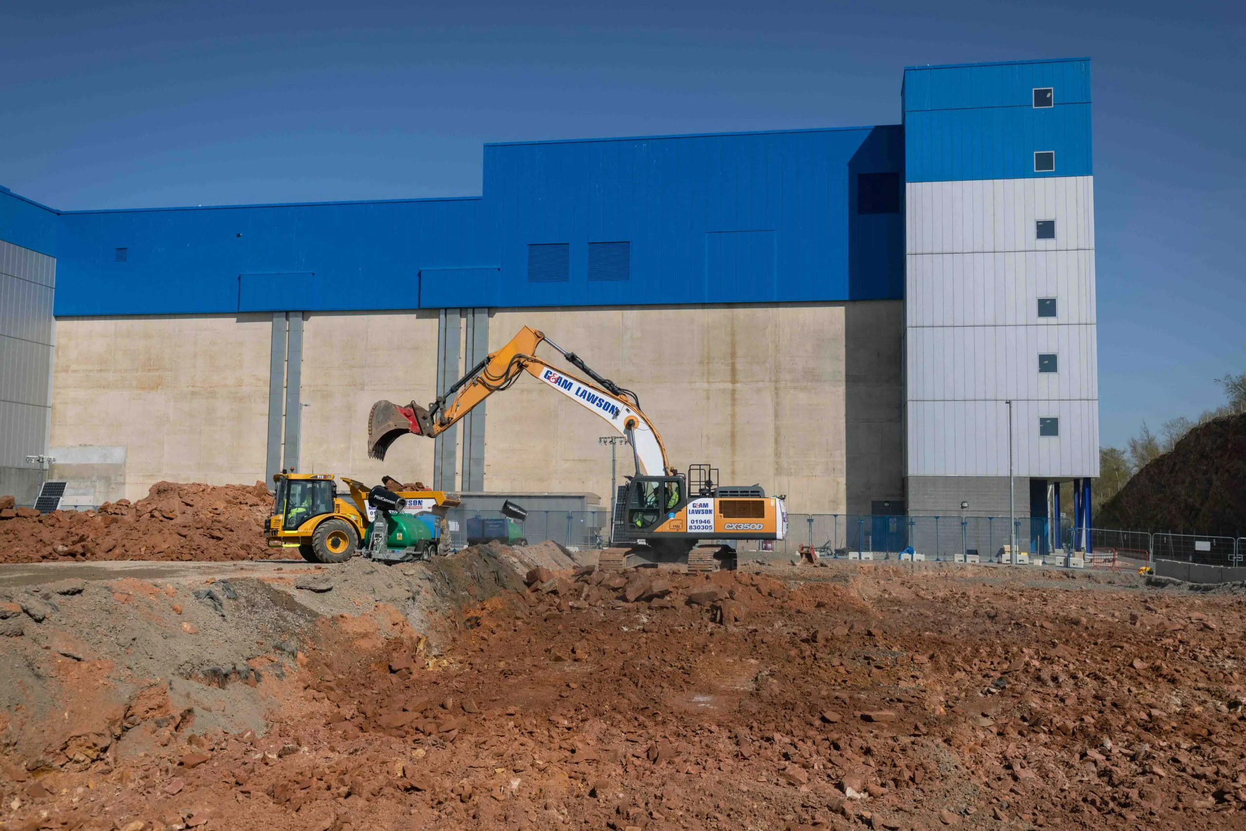 Excavator loads dirt into another machine’s bucket near large industrial buildings painted blue and grey.