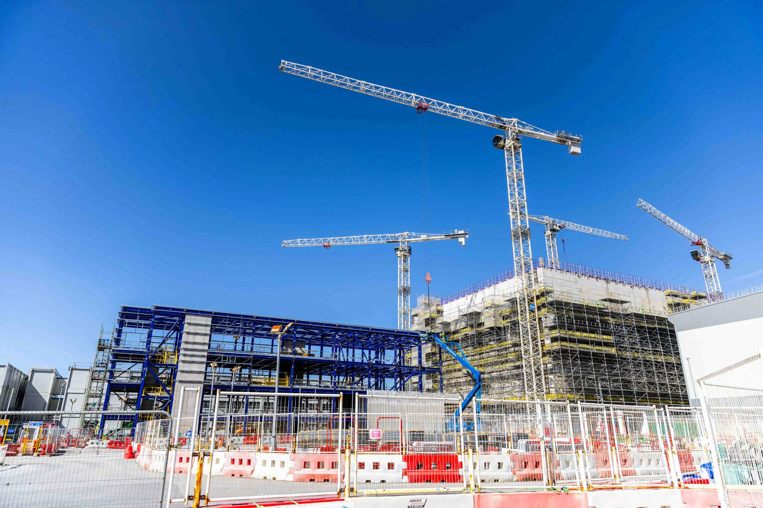 Large construction site with multiple cranes erecting steel structures and safety barriers surrounding the area.