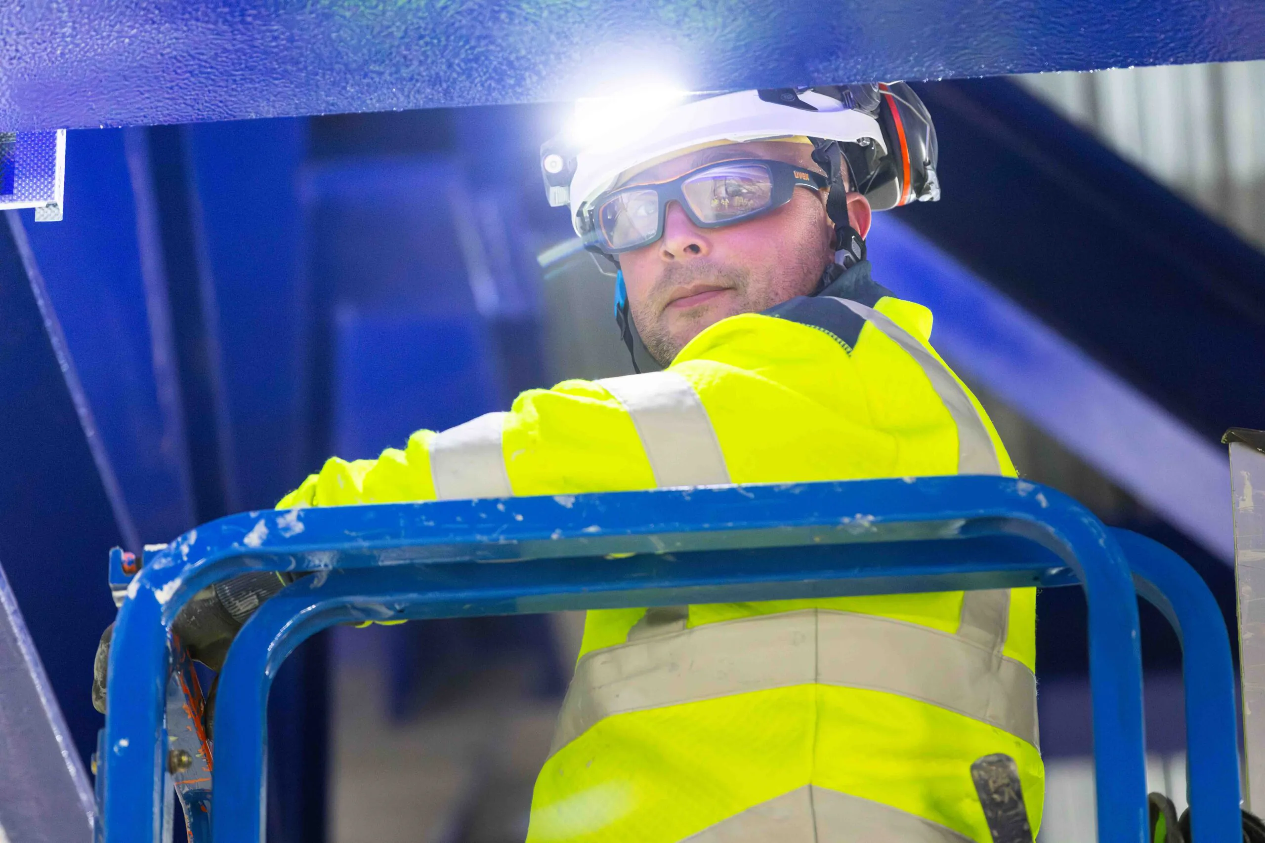 Close-up of a construction worker wearing goggles and helmet on an elevated platform with blue structural elements behind.