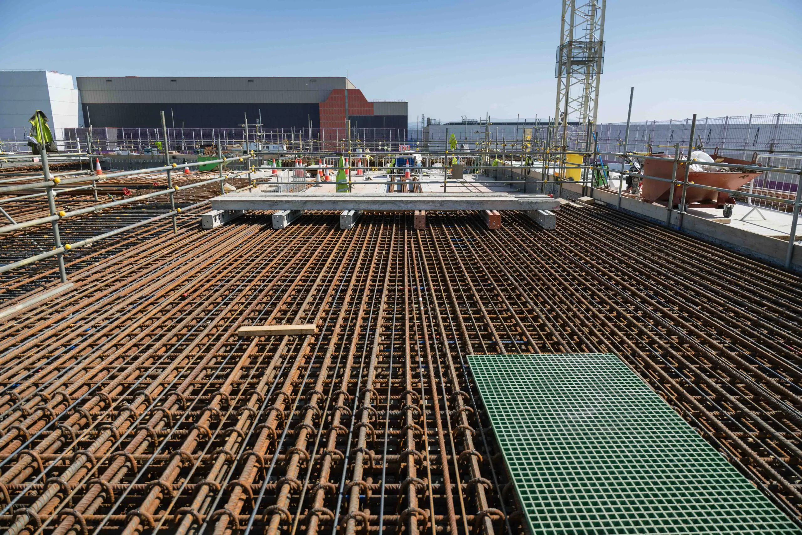 Rebar grid laid out for concrete pouring at a large construction site surrounded by scaffolding and cranes.