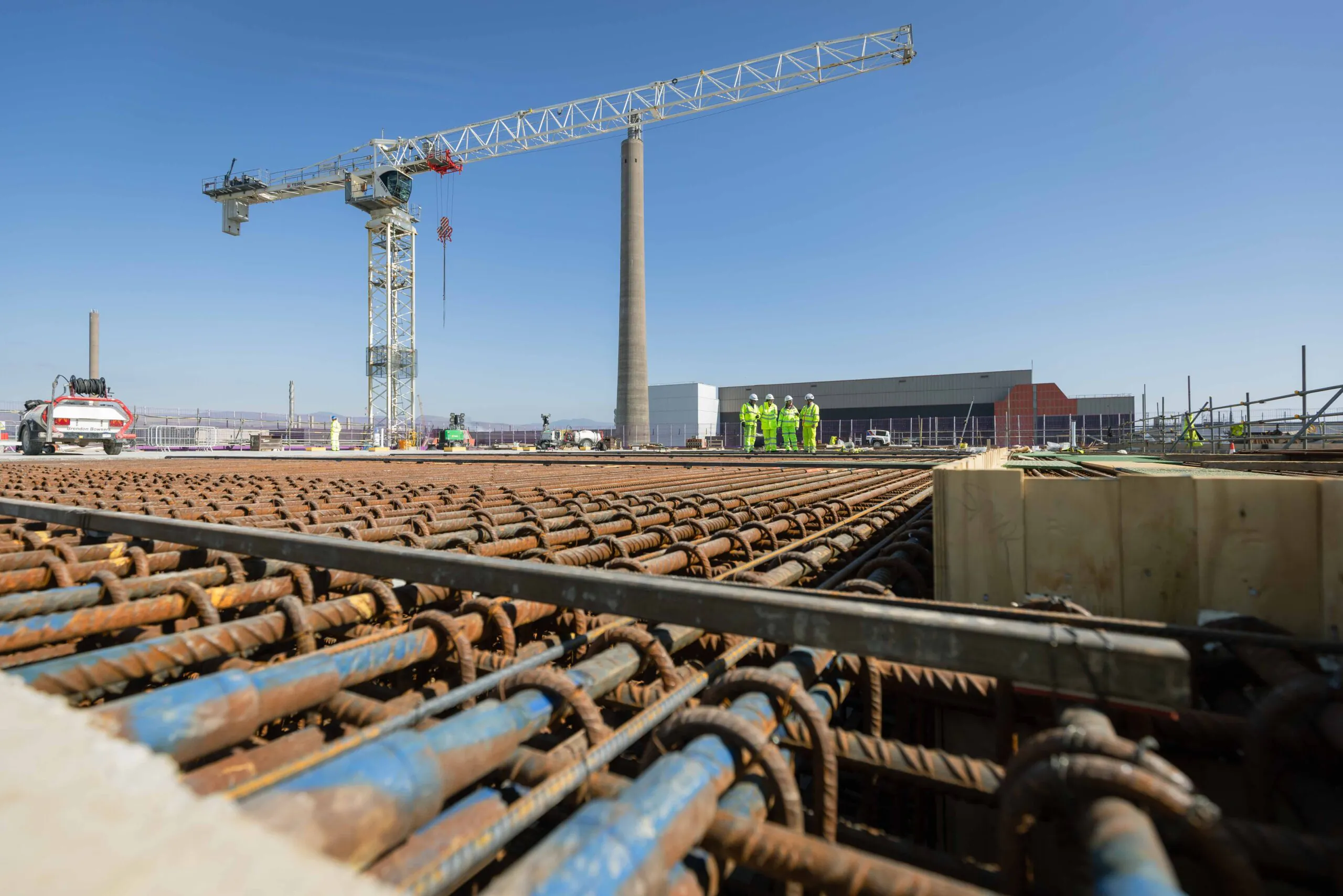 Construction site with rebar laid out for concrete pouring, a large crane in the background, and workers in high-visibility clothing nearby.