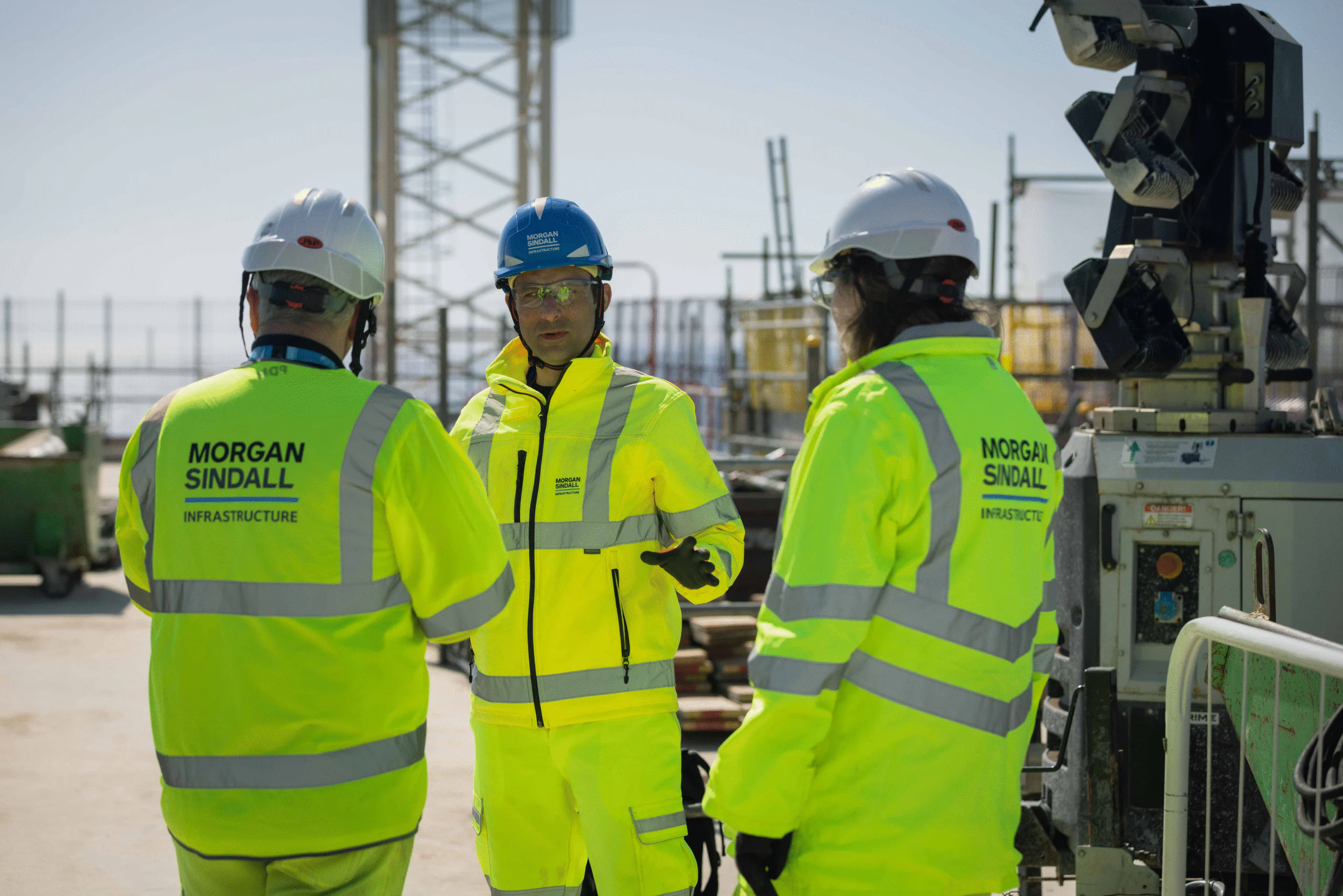 Three construction workers in yellow high-visibility jackets discuss work at an outdoor site; jackets read “MORGAN SINDALL INFRASTRUCTURE.”