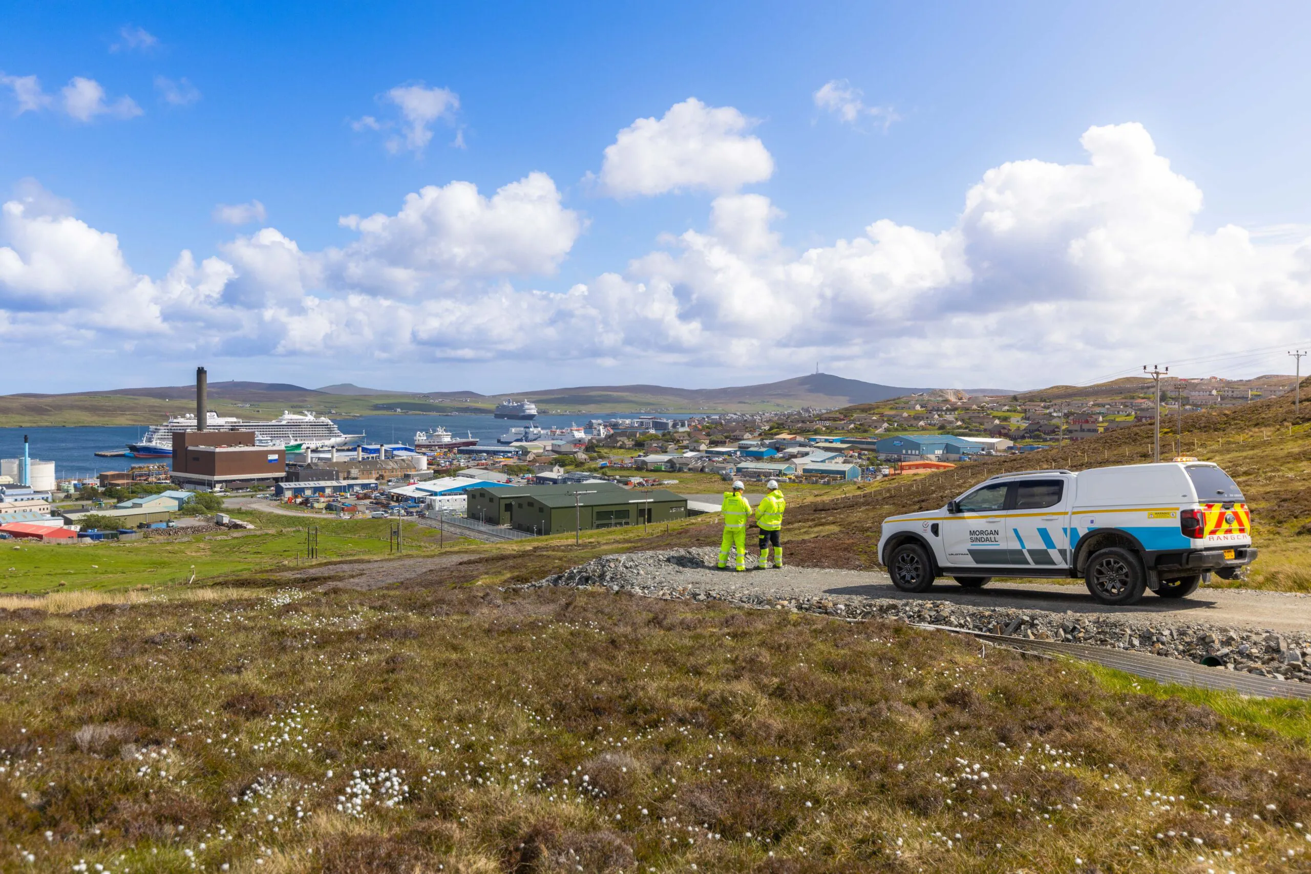 Two workers stand on a gravel path overlooking a coastal townscape with harbor, buildings, and grassy hills; a “Morgan Sindall” utility vehicle is parked nearby.
