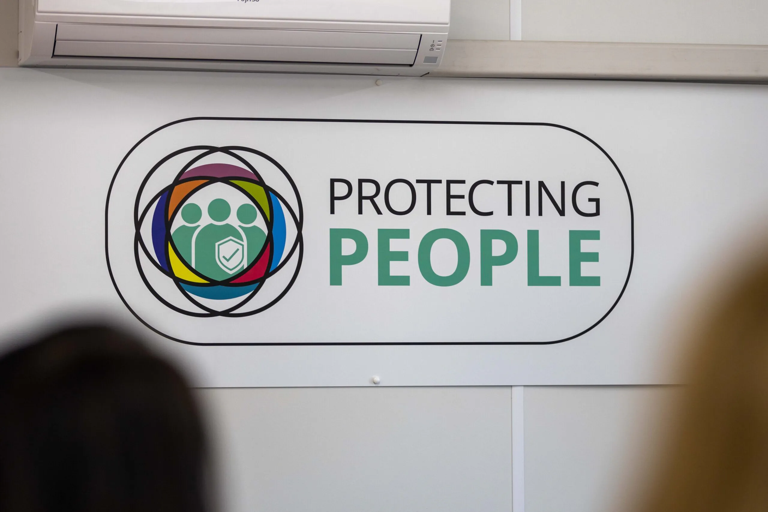 Wall-mounted sign reading “PROTECTING PEOPLE” in bold letters, featuring a logo with three stylized human figures inside overlapping colored circles and a shield. An air conditioning unit is installed above the sign.