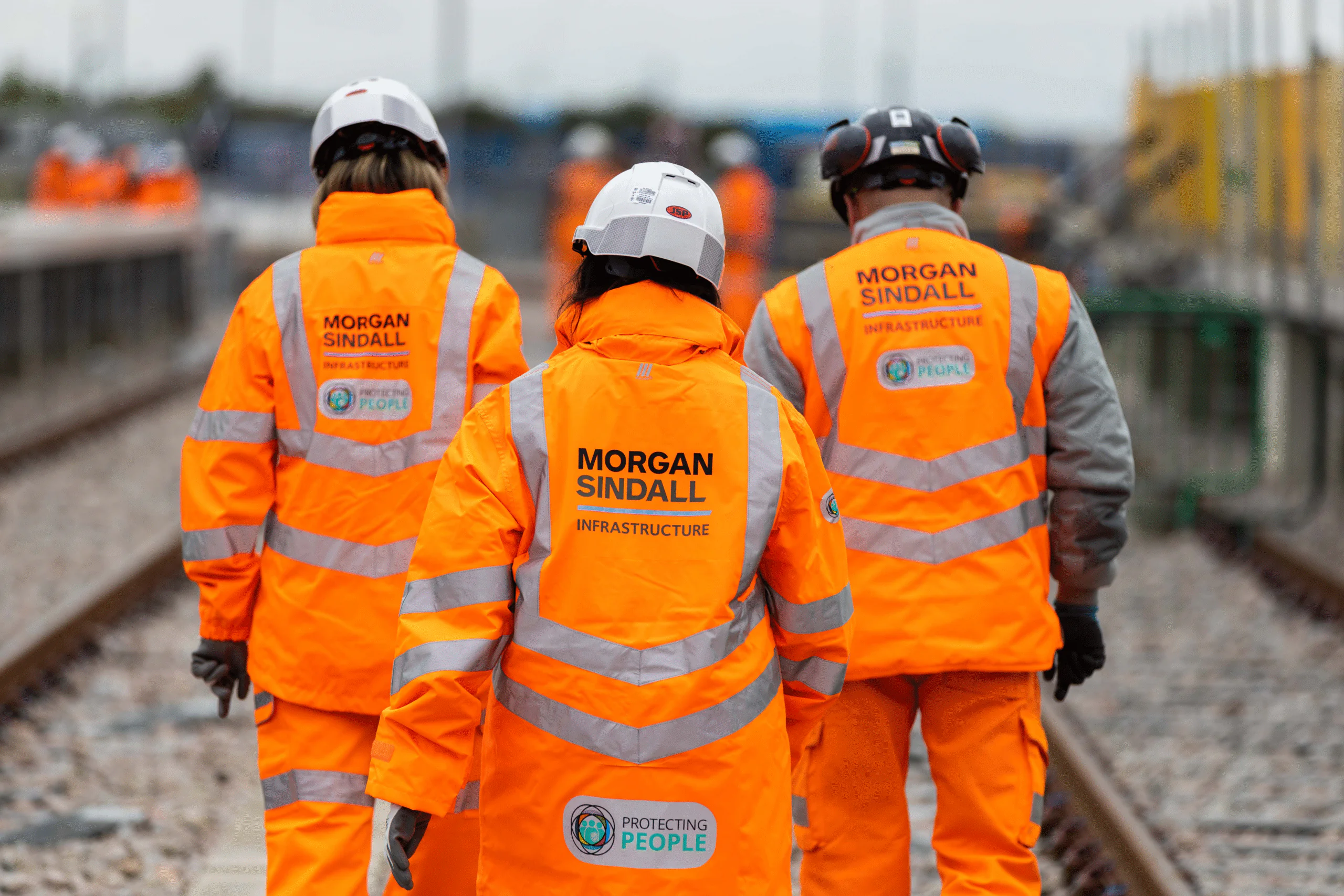Three construction workers in orange safety gear walk along railway tracks with “MORGAN SINDALL INFRASTRUCTURE” printed on their jackets.