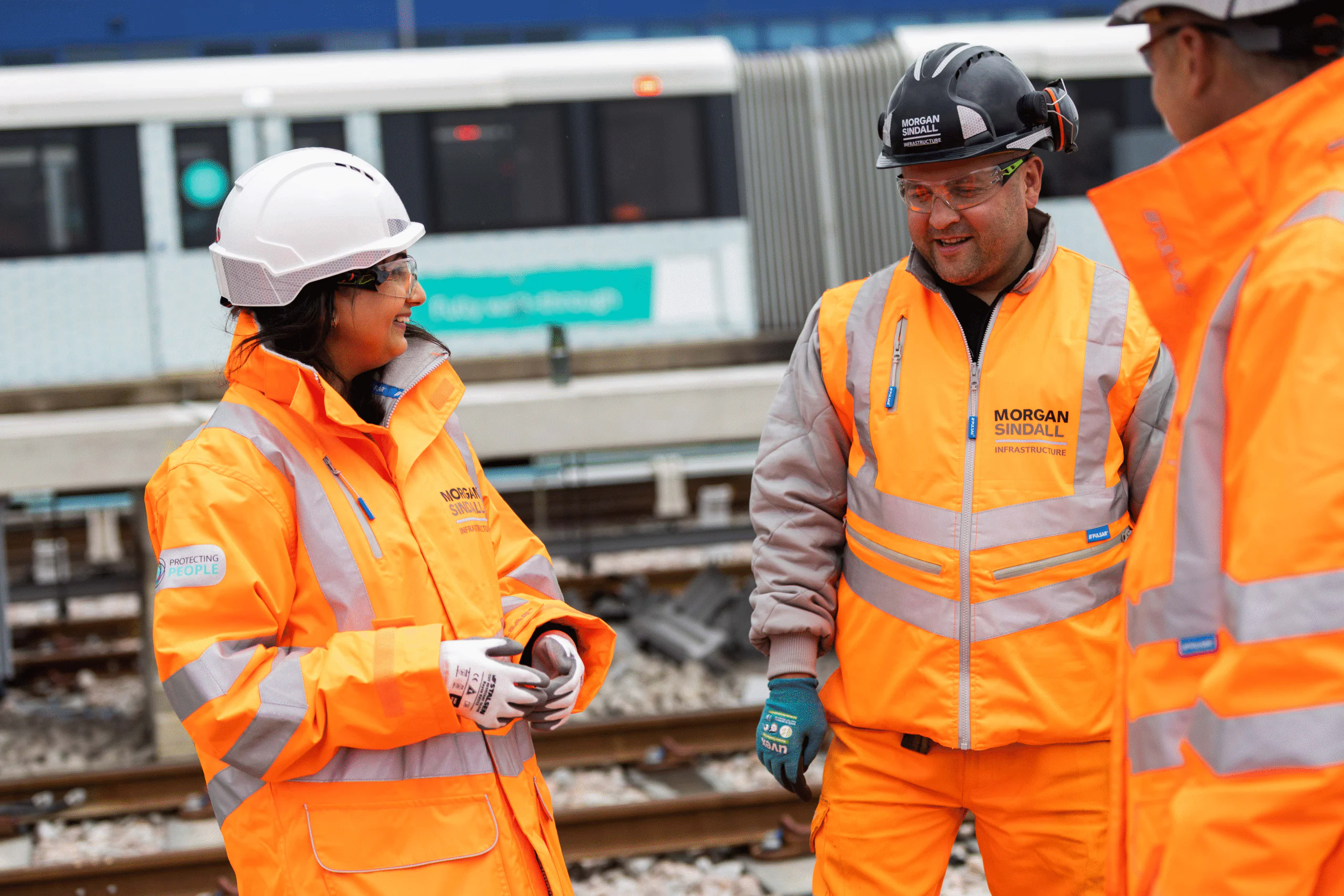 Three workers in orange high-visibility jackets and hard hats converse near railway tracks