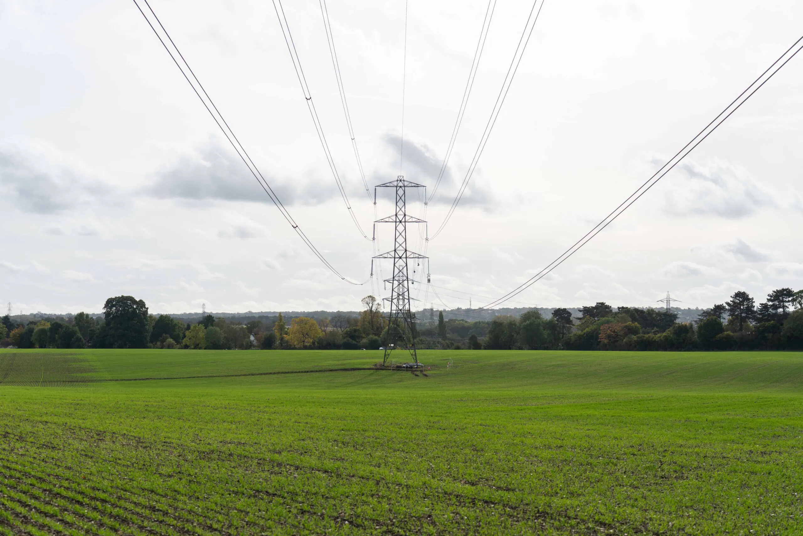 Green field with power lines extending from a tall metal lattice tower under an overcast sky.