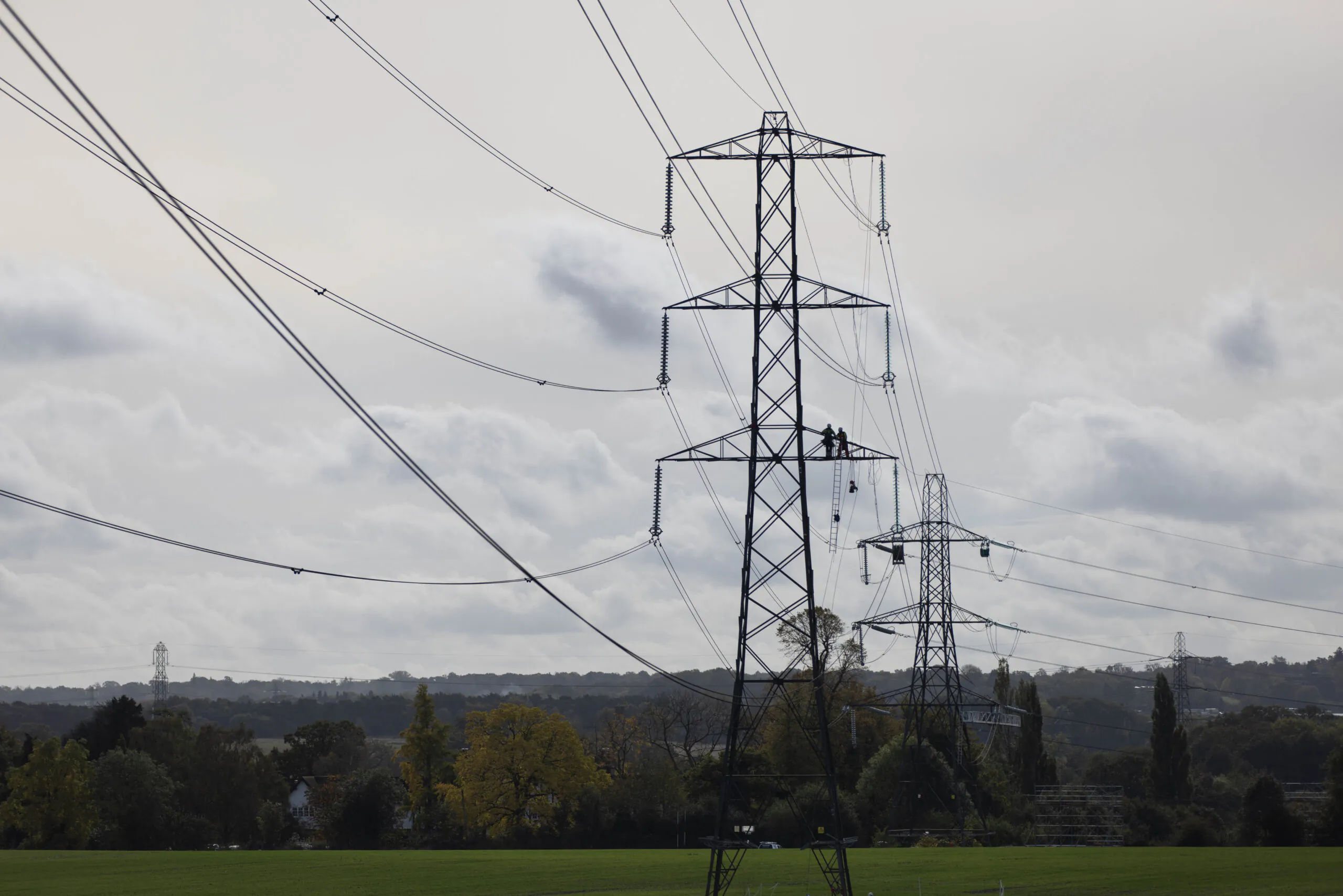 Workers in safety gear inspect and work on transmission towers and power lines in a rural area.