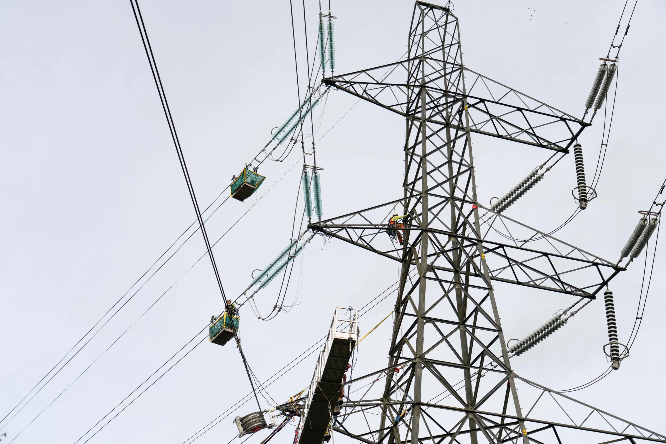 Workers suspended from cables perform maintenance on high-voltage power lines connected to tall transmission towers under cloudy skies.