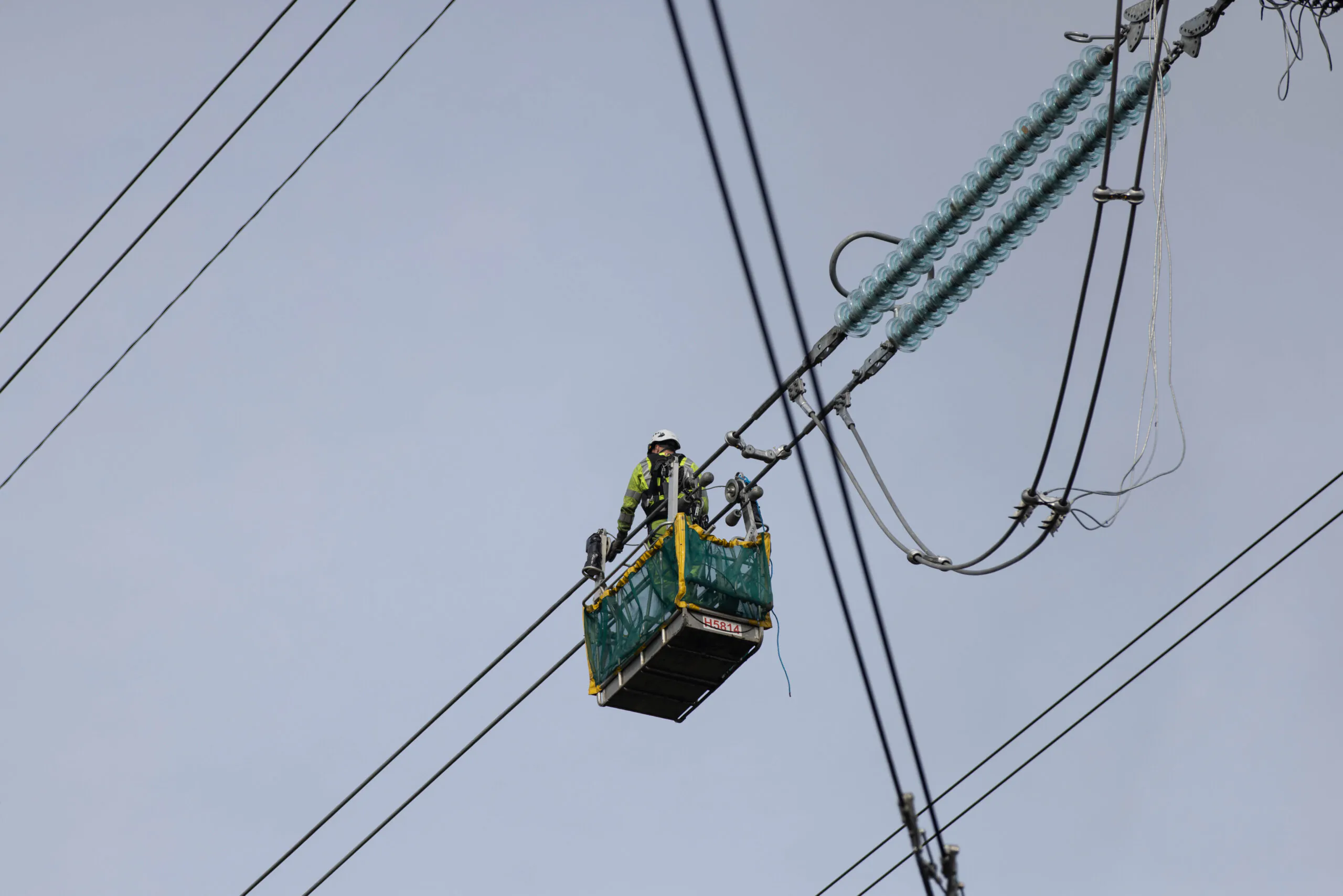 Worker performing maintenance inside an elevated basket attached to power lines high above ground level against grey skies.