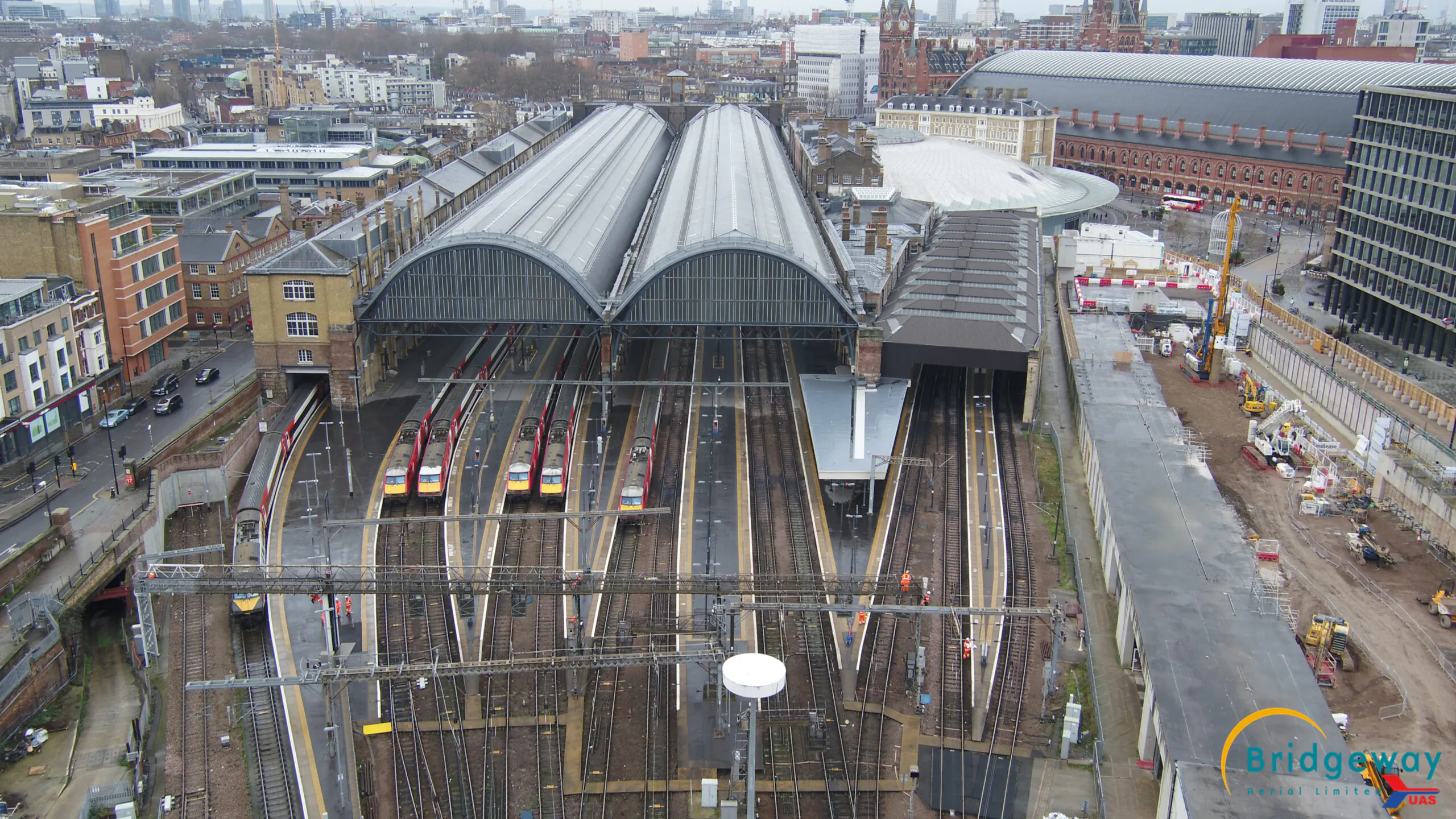Aerial view of a train station with multiple tracks leading into two large arched-roof buildings, surrounded by trains, buildings, and construction areas. Bridgeway logo in the bottom right corner.