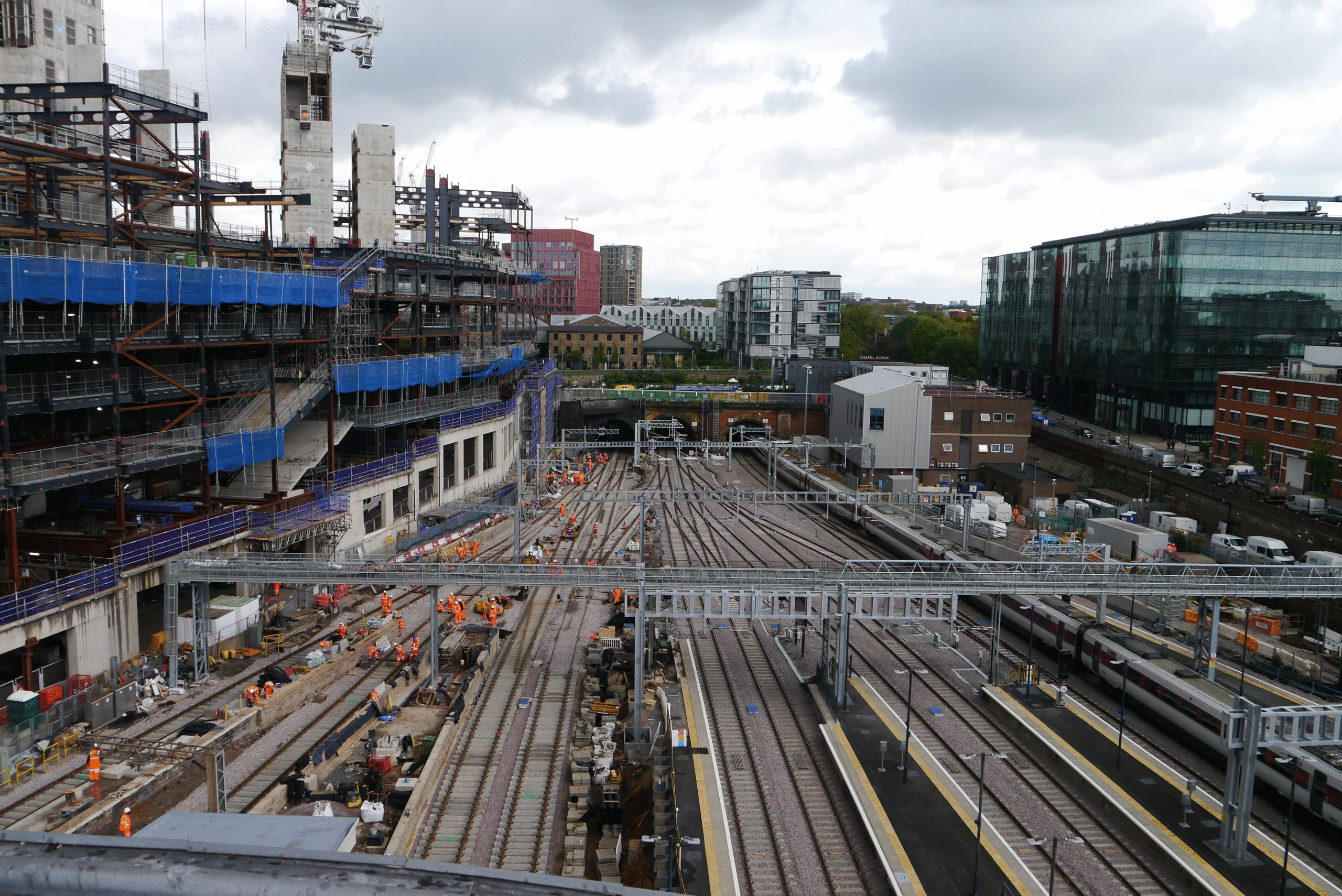 Railway station construction site with multiple tracks and platforms under development. Workers in orange safety gear, scaffolding, cranes, and construction materials are visible, with modern buildings in the background.