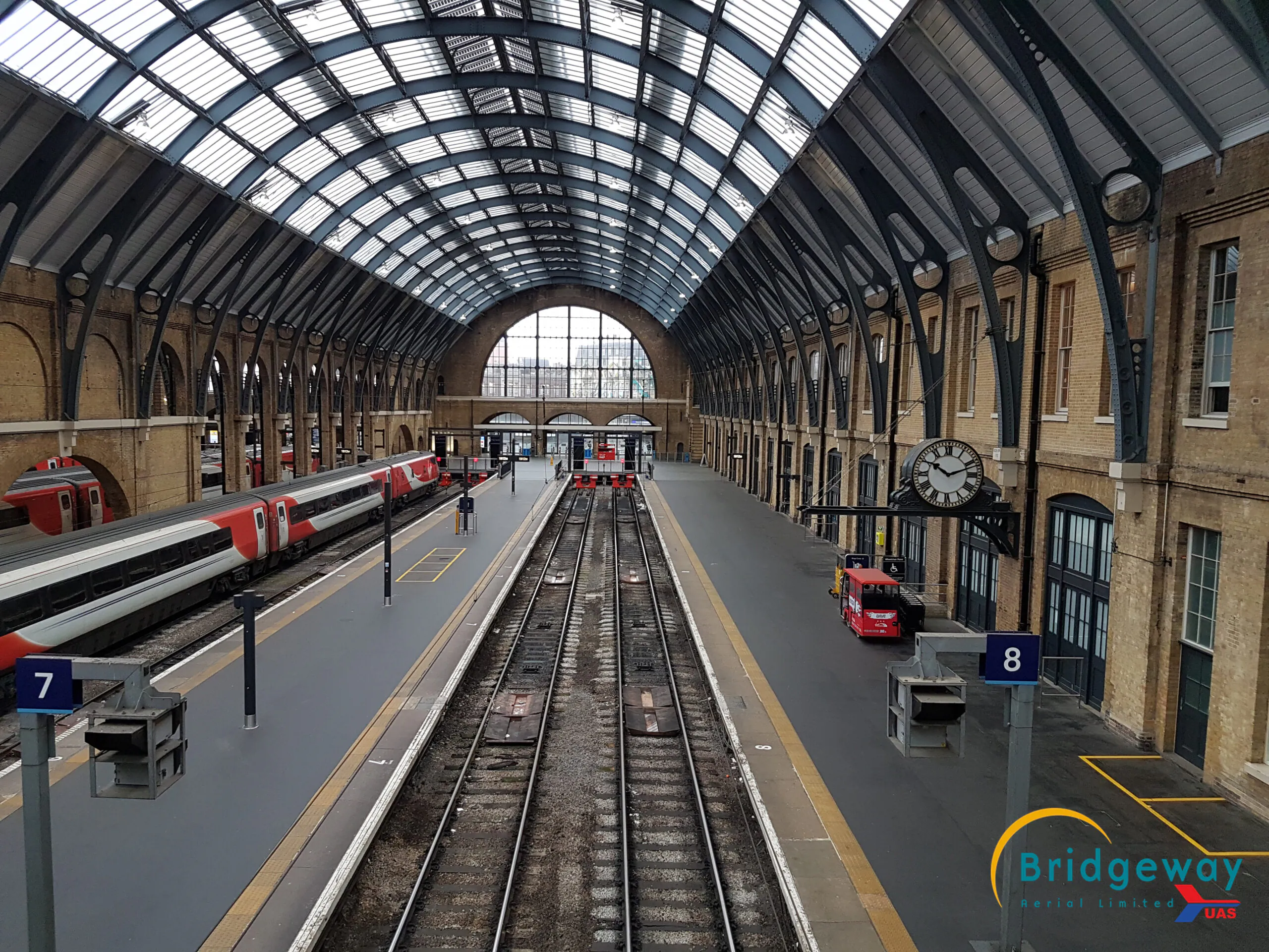 Interior view of a large train station with multiple platforms and tracks under a high arched glass roof. Platform numbers 7 and 8 are visible, along with a clock and trains on the tracks. Bridgeway logo in the bottom right corner.
