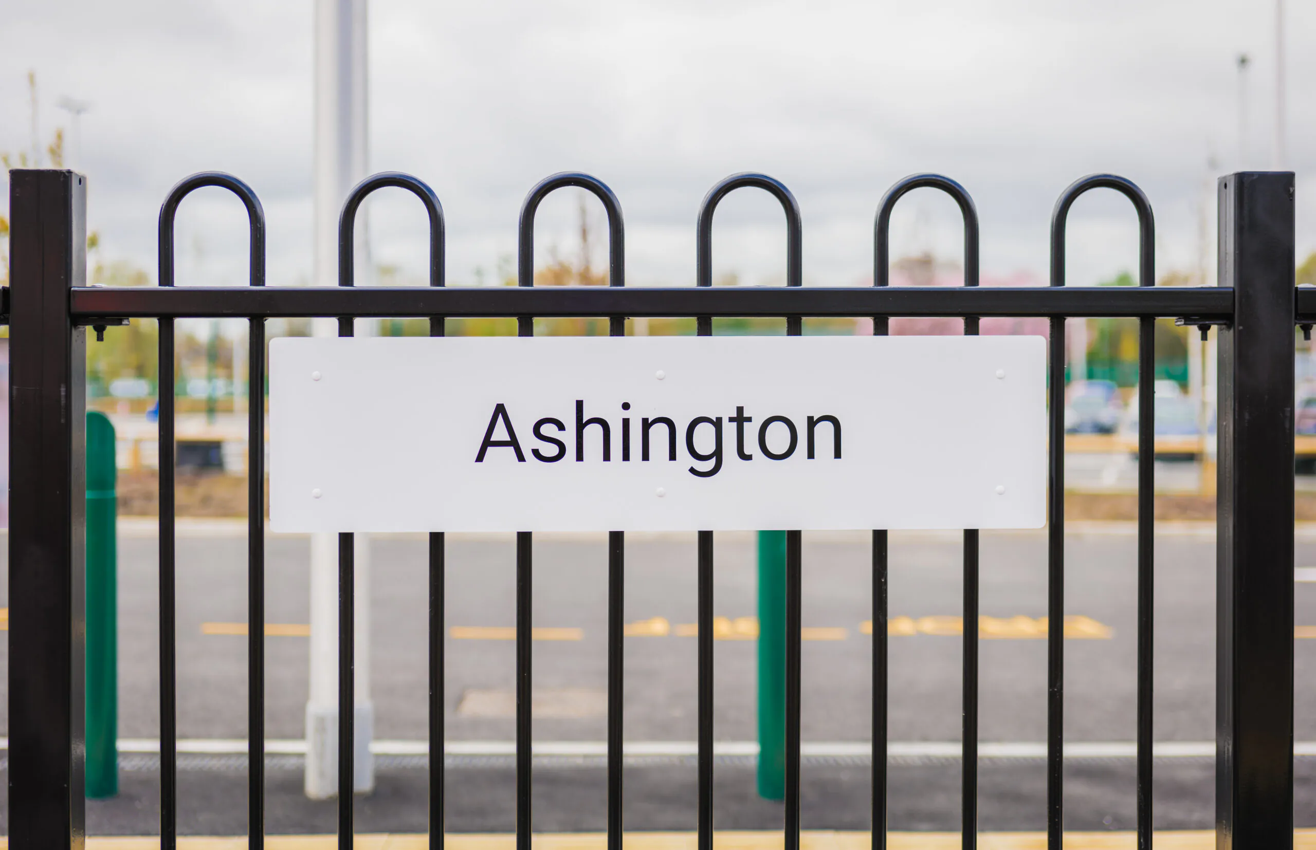White 'Ashington' sign mounted on a black fence with parking spaces visible behind it.
