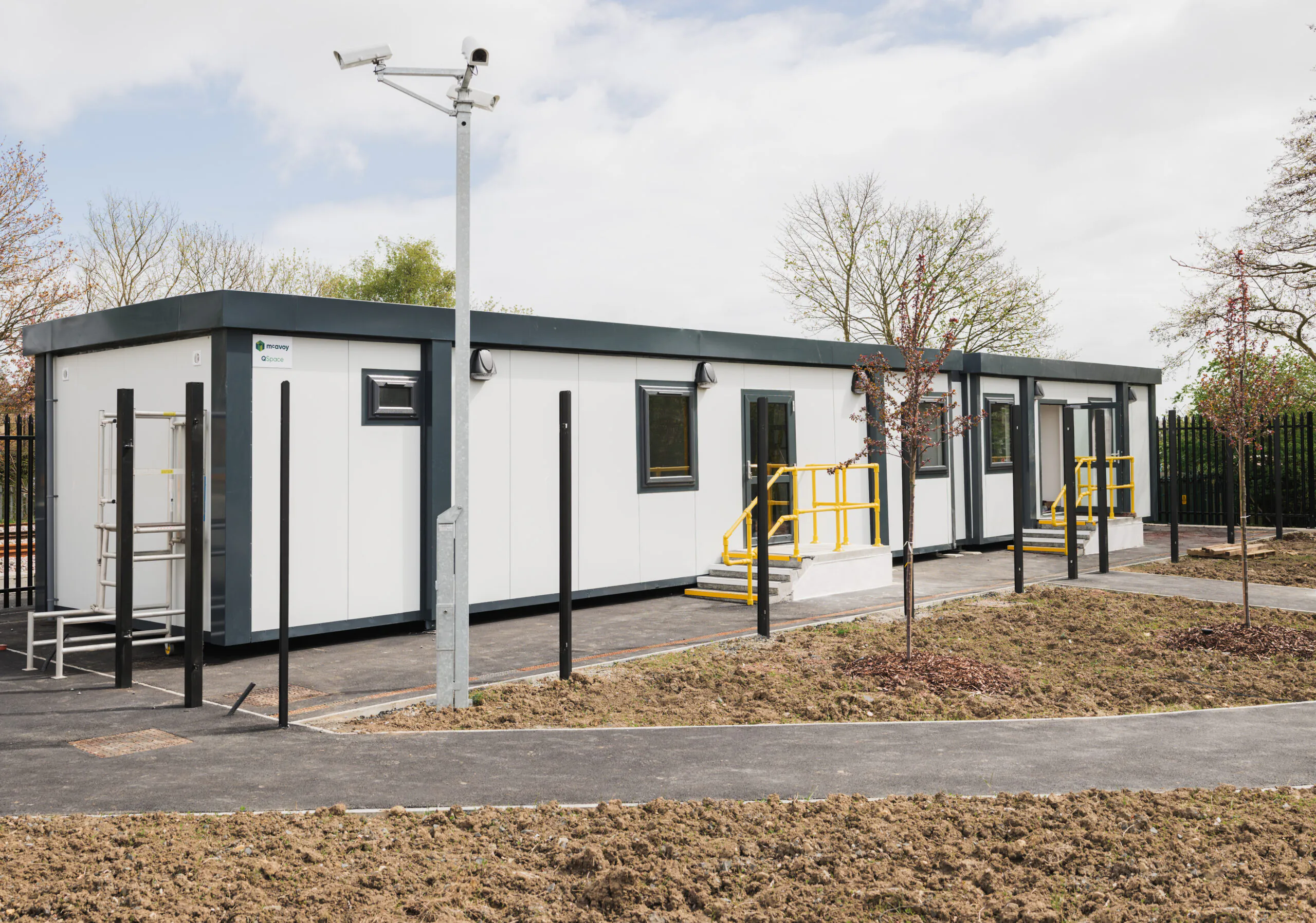 Small modular building surrounded by newly planted trees and pathways within a fenced area.