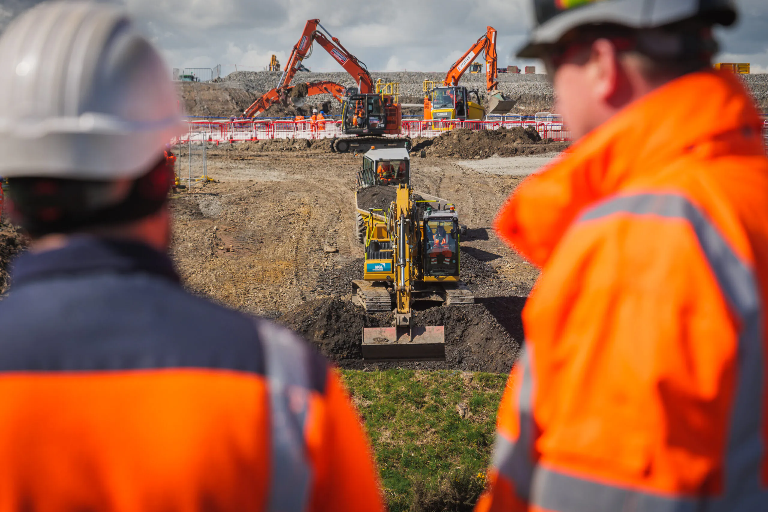 Two people in high-visibility clothing observing a construction site with multiple excavators in operation.