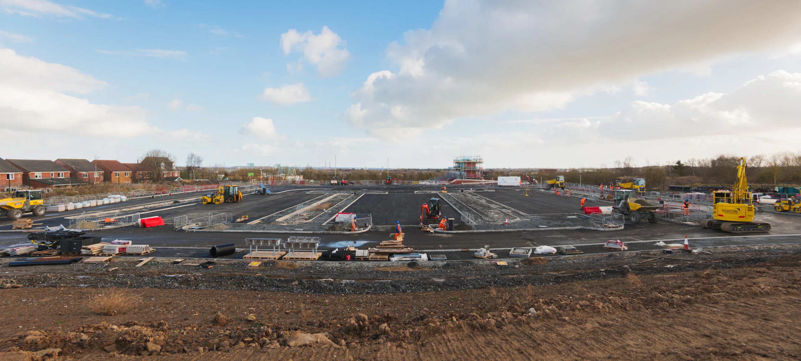 Wide-angle view of a construction site with excavators and bulldozers operating near residential houses under a partly cloudy sky."