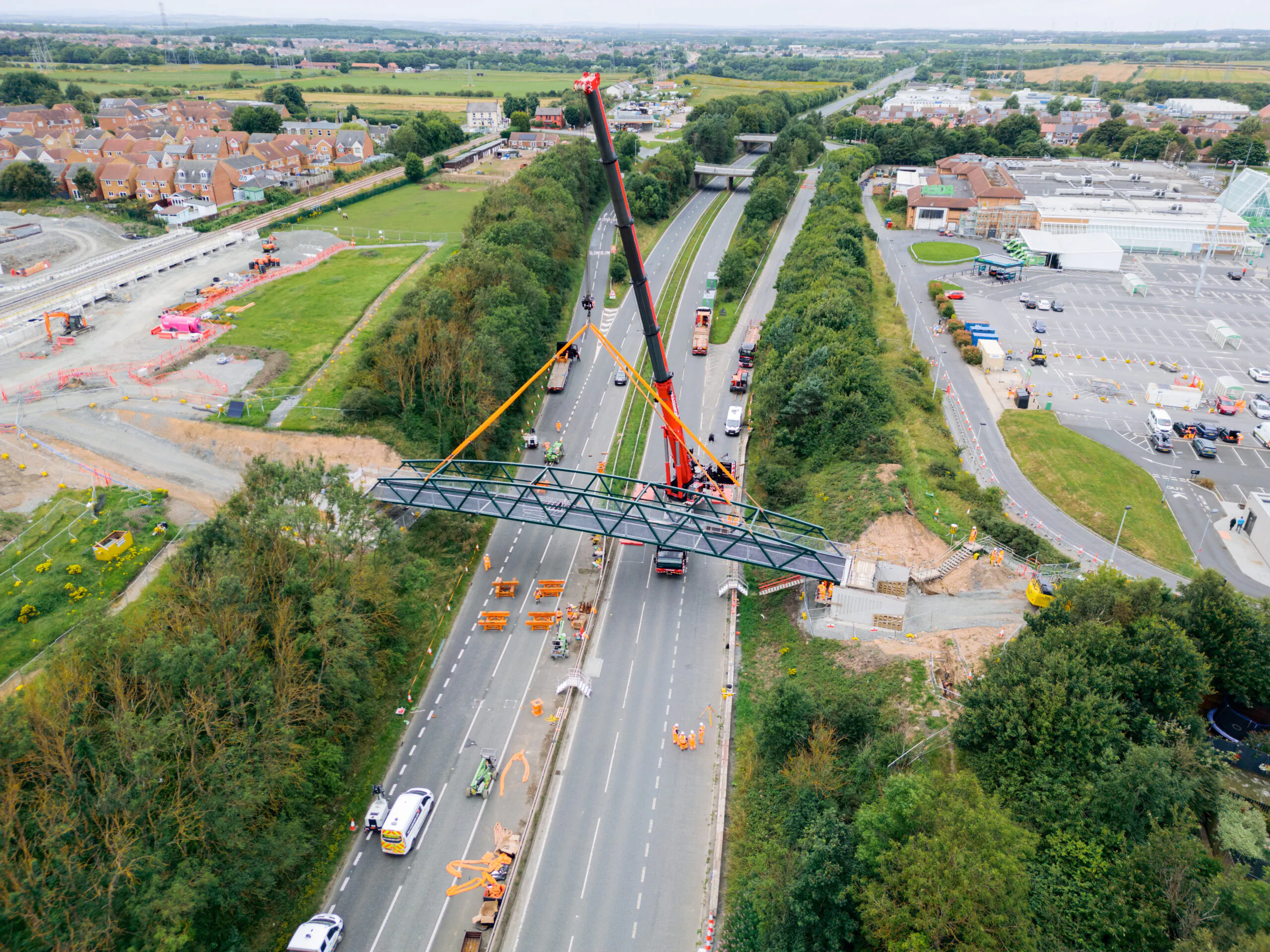 Aerial view of a crane lifting a steel structure over multiple lanes of traffic near residential and commercial areas.