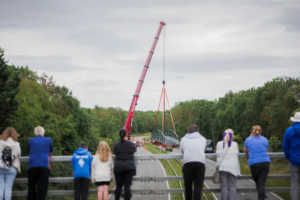 People standing on an overpass watching a crane lift a large steel structure over a road surrounded by trees.