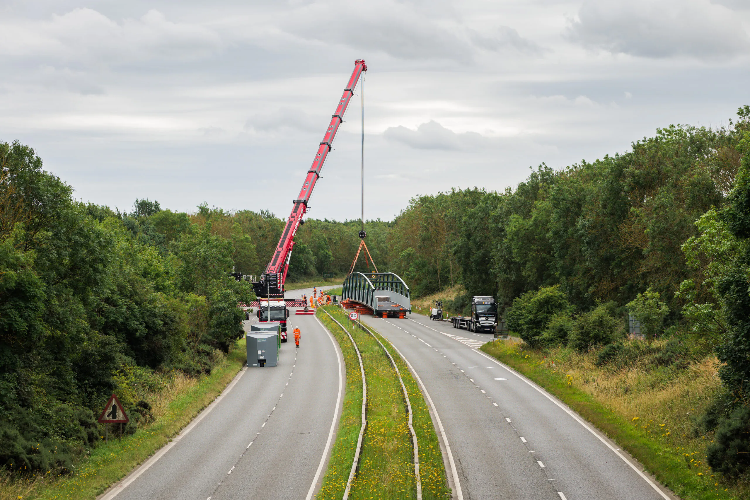 Workers using cranes to lift large steel structures onto flatbed trucks along a divided highway bordered by forest.