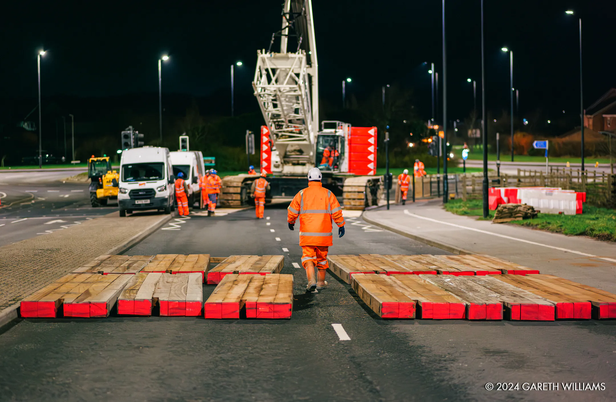 Workers assembling crane components at a construction site with lighting and safety barriers in place.