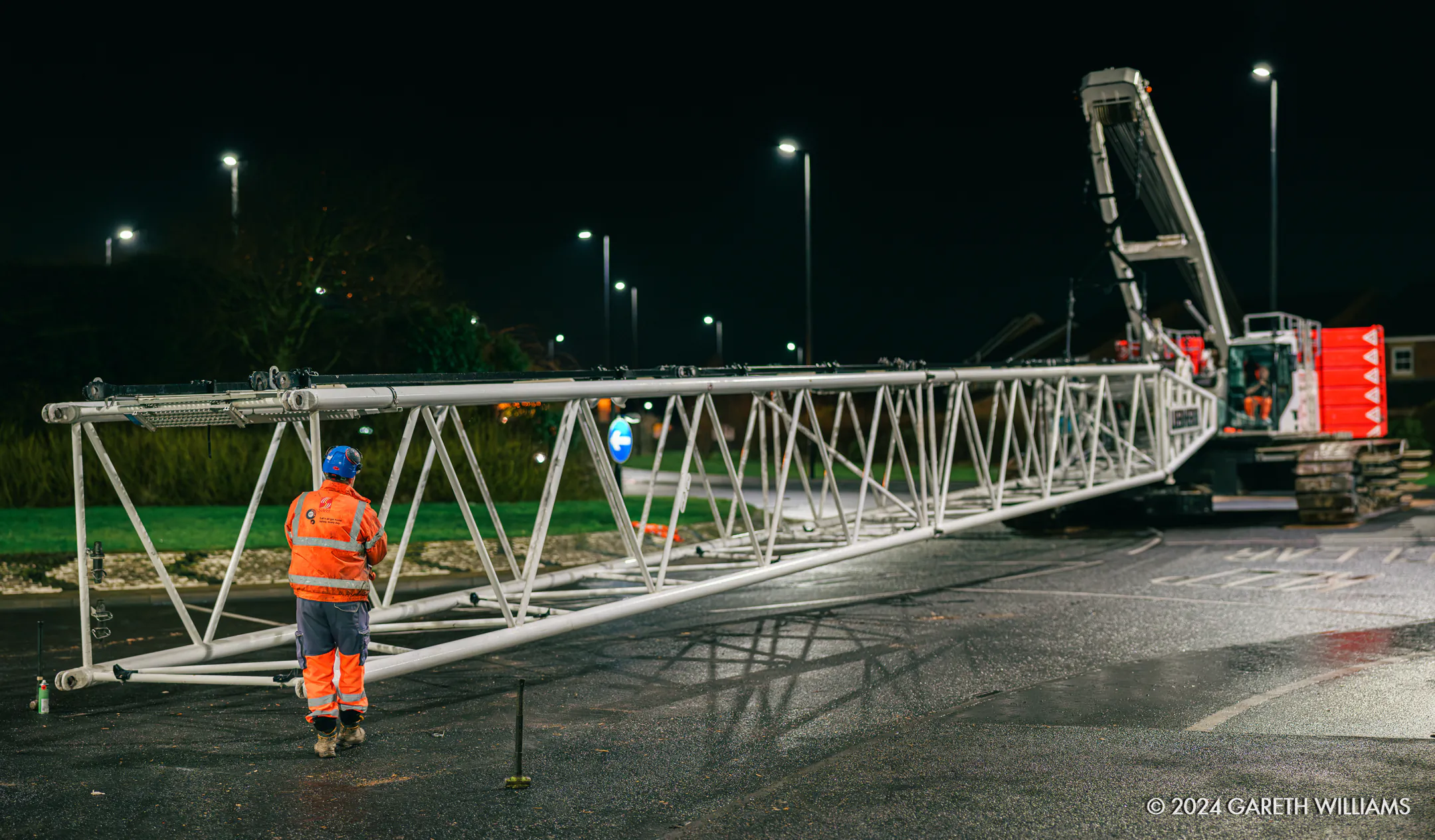 Nighttime scene of workers assembling or disassembling crane components under artificial lighting in an open area.