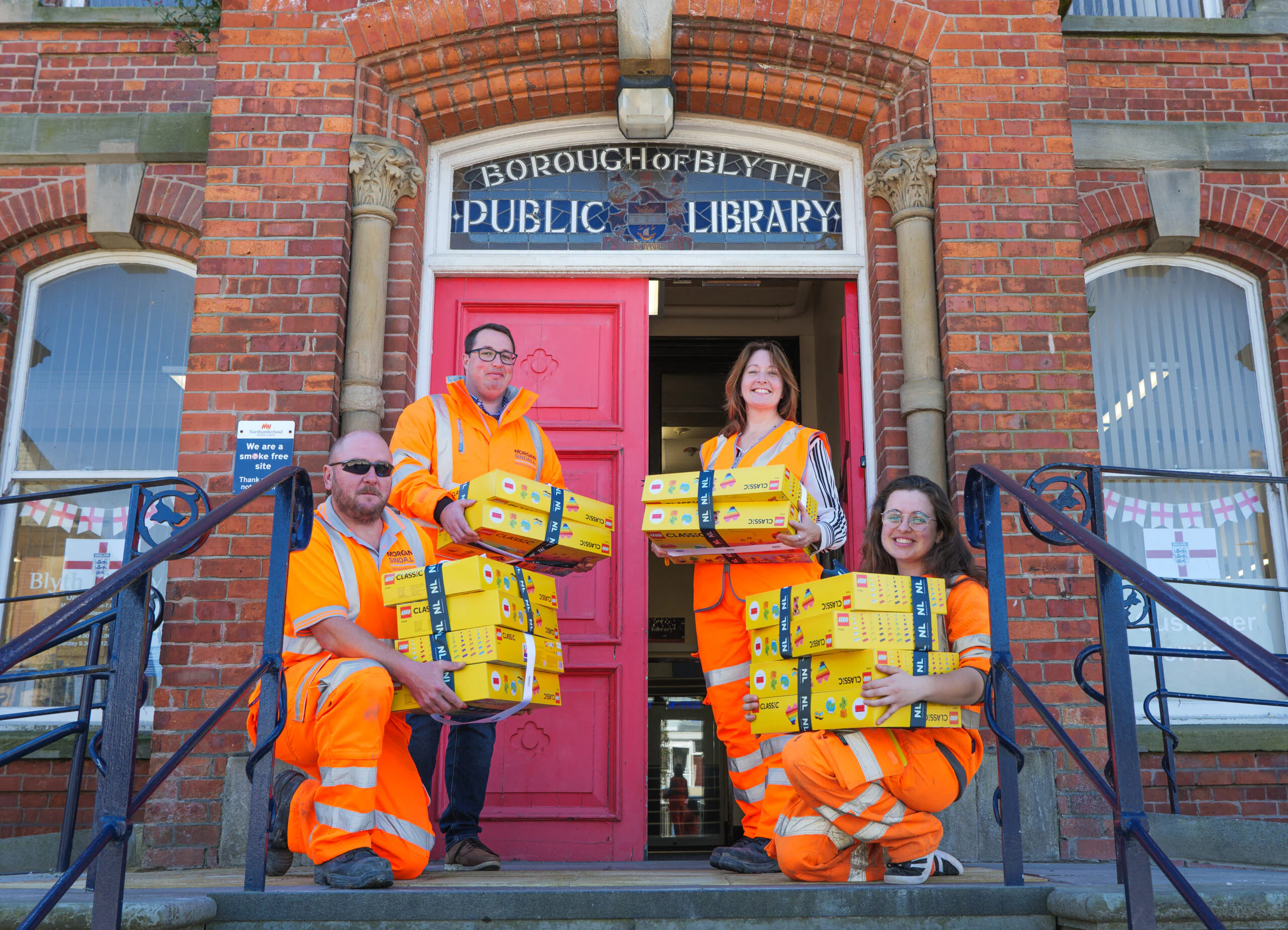 Four individuals in bright orange safety uniforms are positioned on the steps of the Blyth Public Library, a brick building with a red door and arched windows. Each person is holding or carrying yellow boxes labeled "Jiffy," suggesting a delivery or collection event. A sign above the door reads "Borough of Blyth Public Library.