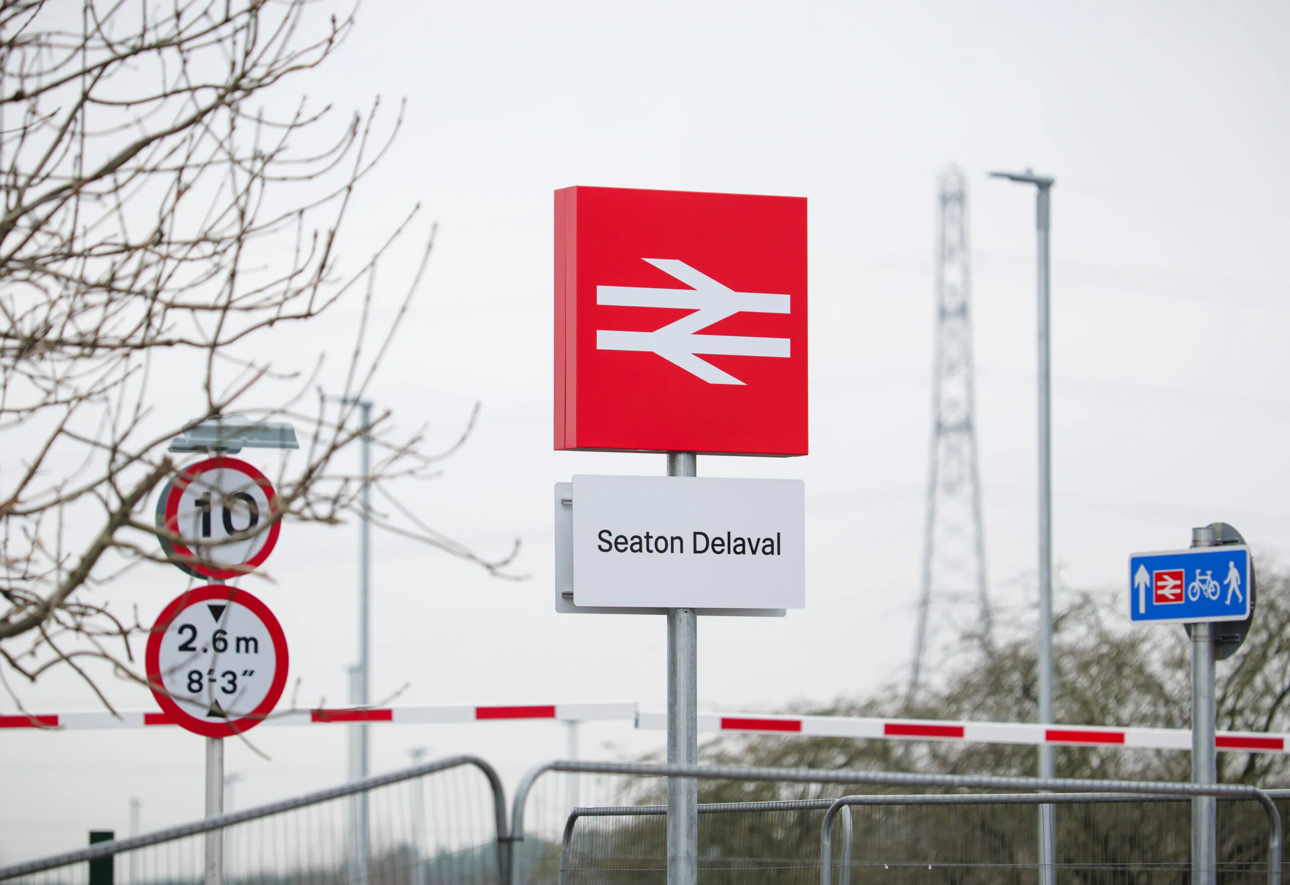 Railway signage at Seaton Delaval station showing the British Rail double arrow logo above the station name. Nearby signs include a 10 mph speed limit, a 2.6m (8'6") height restriction, and a blue pedestrian and cycle path sign. The area is fenced, with leafless trees and utility poles in the background.