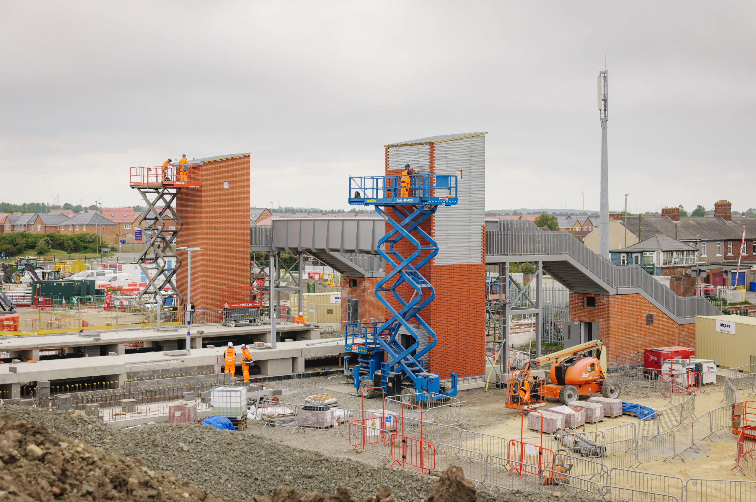 Daytime construction scene with two scissor lifts and workers building brick structures connected by metal walkways.