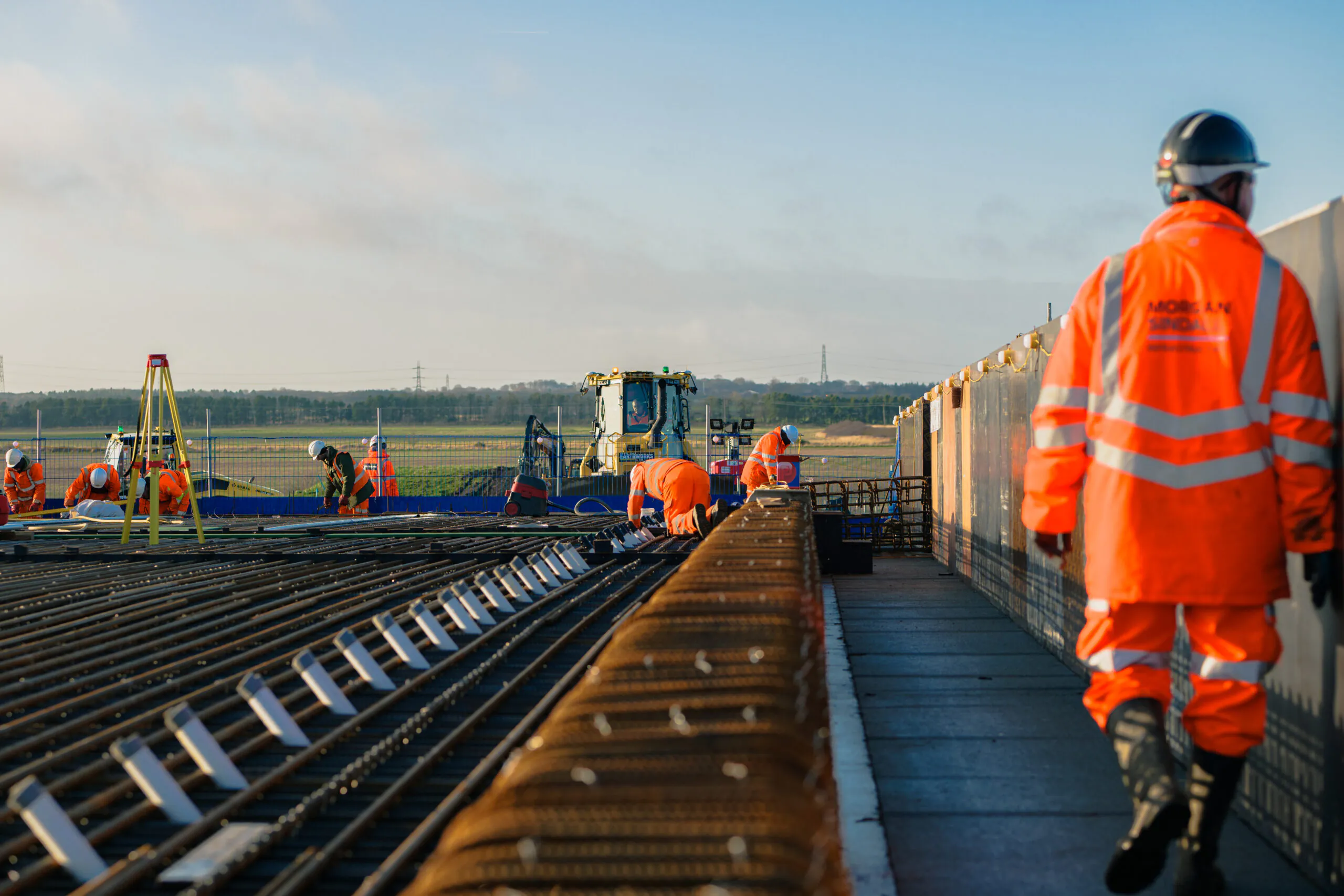 Workers in high-visibility clothing working on rebar at an outdoor construction site with machinery in the background.